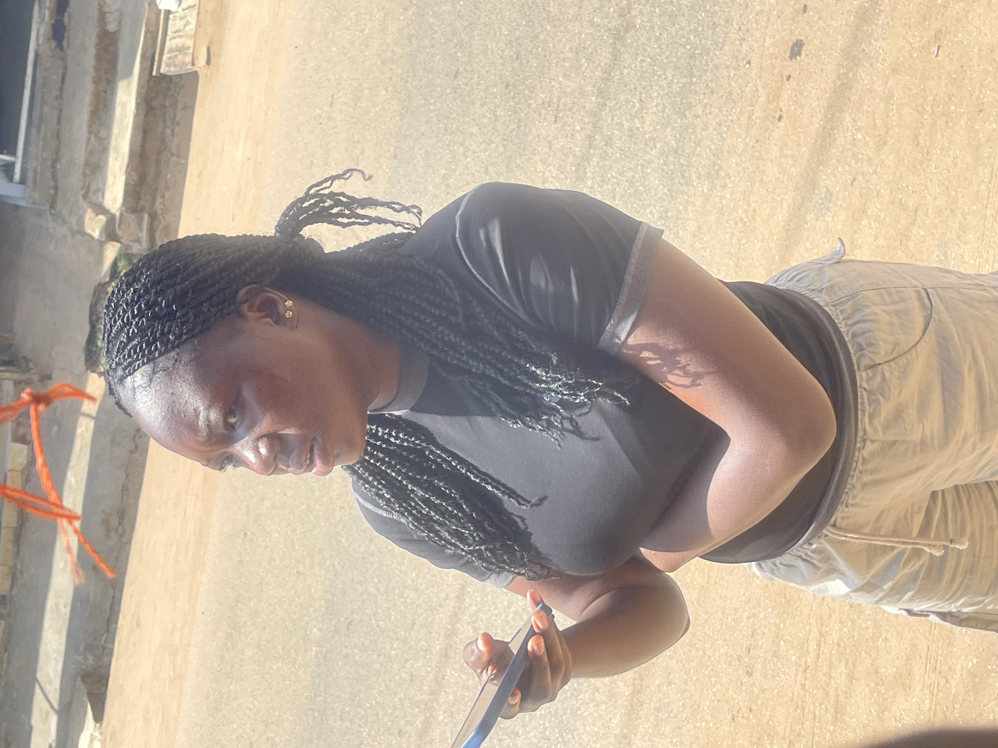 A medium shot captures a young Black woman with braided hair, wearing a black t-shirt and light-colored shorts, standing outdoors on a paved surface that appears to be a street in Abuja, Nigeria. She is holding a blue smartphone in her left hand and has her arms crossed. The background shows a sun-drenched environment with some concrete structures and orange construction material in the upper left portion of the frame. The lighting suggests it is daytime.
