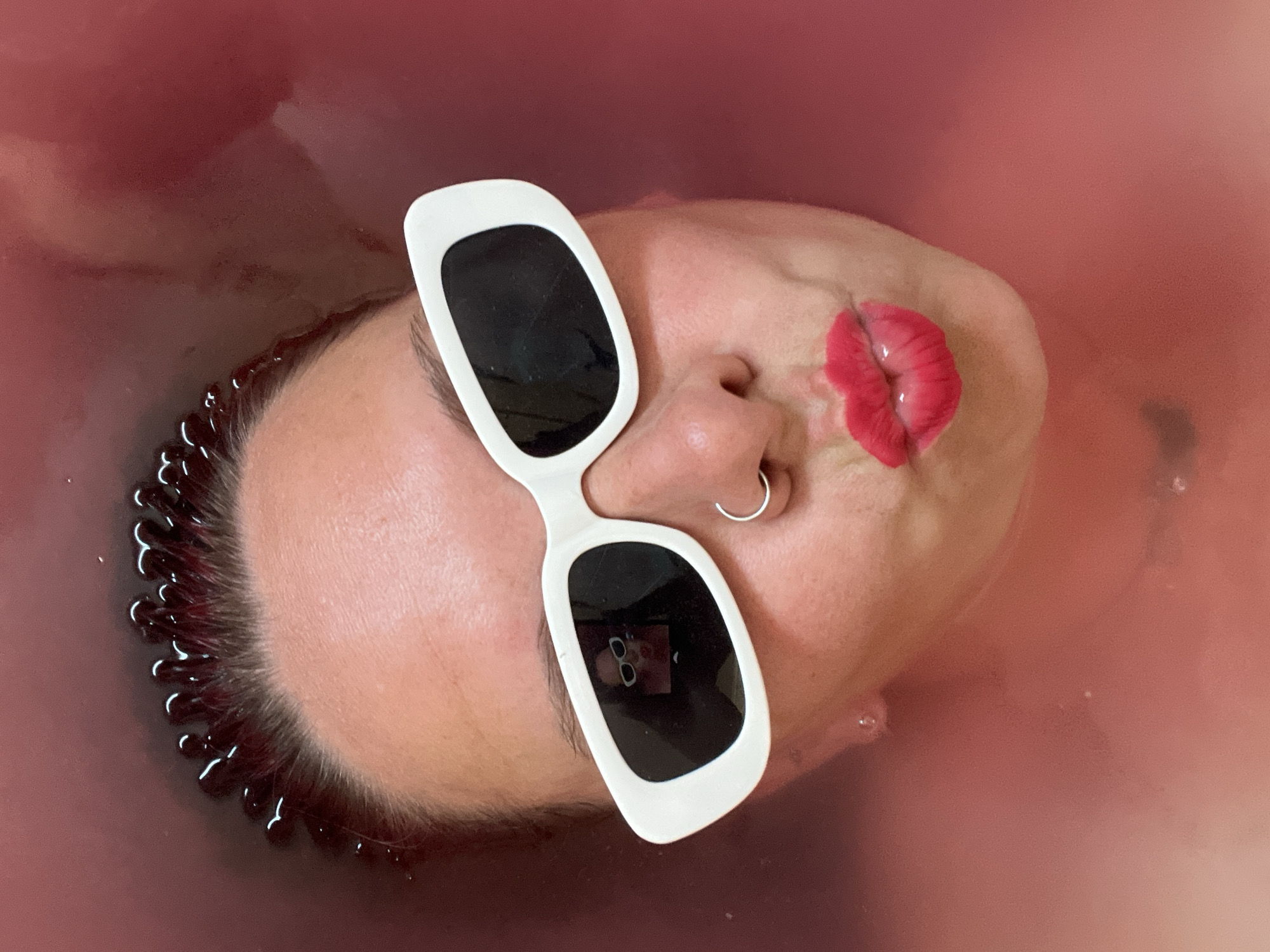 A person is partially submerged in a reddish-pink liquid, possibly water or a bath. The person is wearing white-rimmed sunglasses with black lenses and has a silver nose ring. A pair of bright red lips is positioned next to the person's nose. The edges of the person's hair are visible, along with small droplets clinging to the hair and surrounding liquid. The reflection of sunglasses can be seen in the sunglasses. The exact location in Chaikovsky, Russia, cannot be determined from this image.