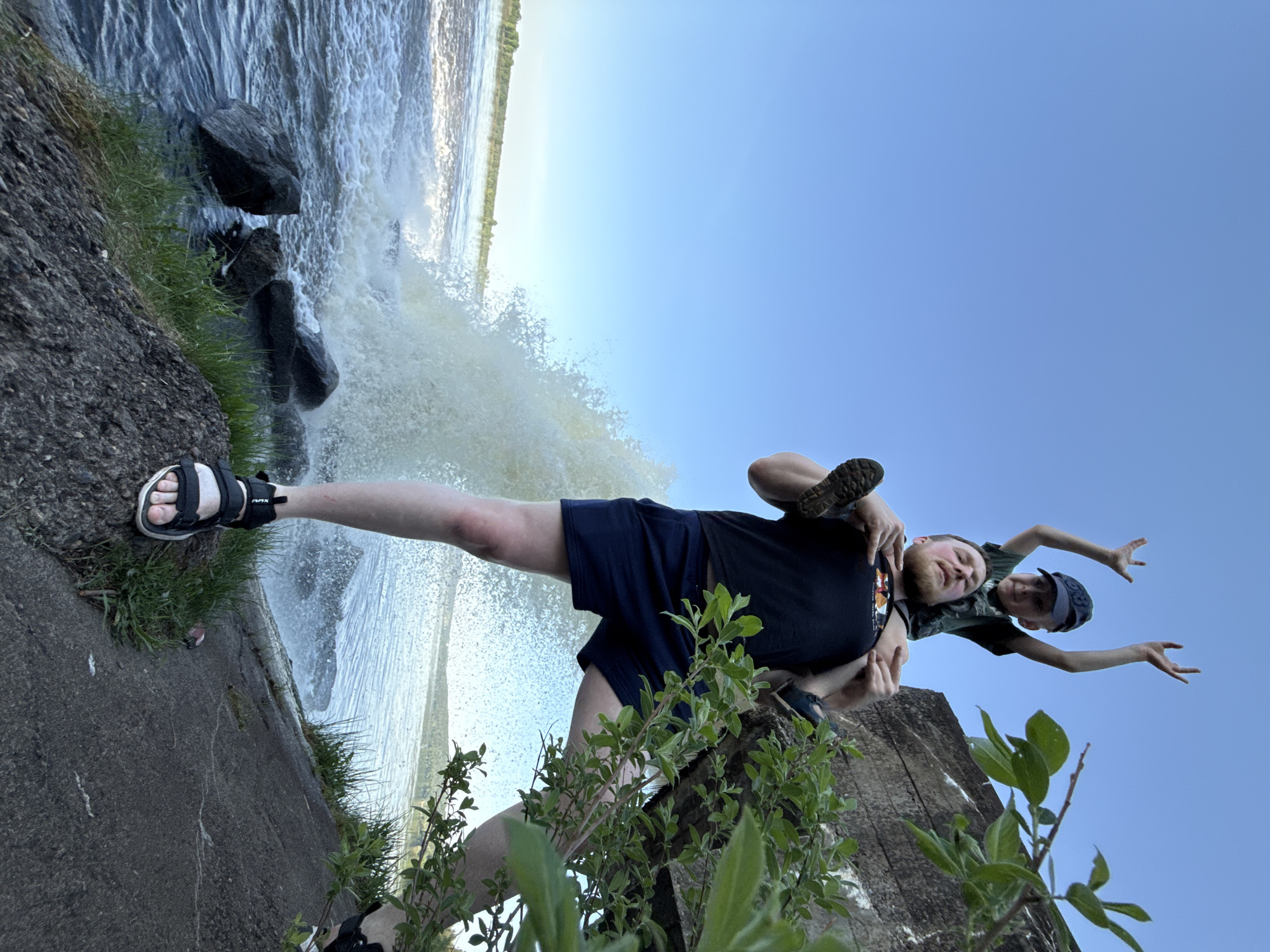 A photograph, rotated counter-clockwise, depicts a man giving a child a piggyback ride against a backdrop of a waterfall and a clear blue sky. The man wears a dark t-shirt, dark shorts, and sandals. The child wears a camouflage shirt and a baseball cap. They both appear to be smiling. The man's left leg is extended toward the left side of the frame, near the edge of the waterfall. Rocks and vegetation are visible near the waterfall's edge. A concrete structure is visible beneath them. The scene is identified as taking place in Novyi, Russia.