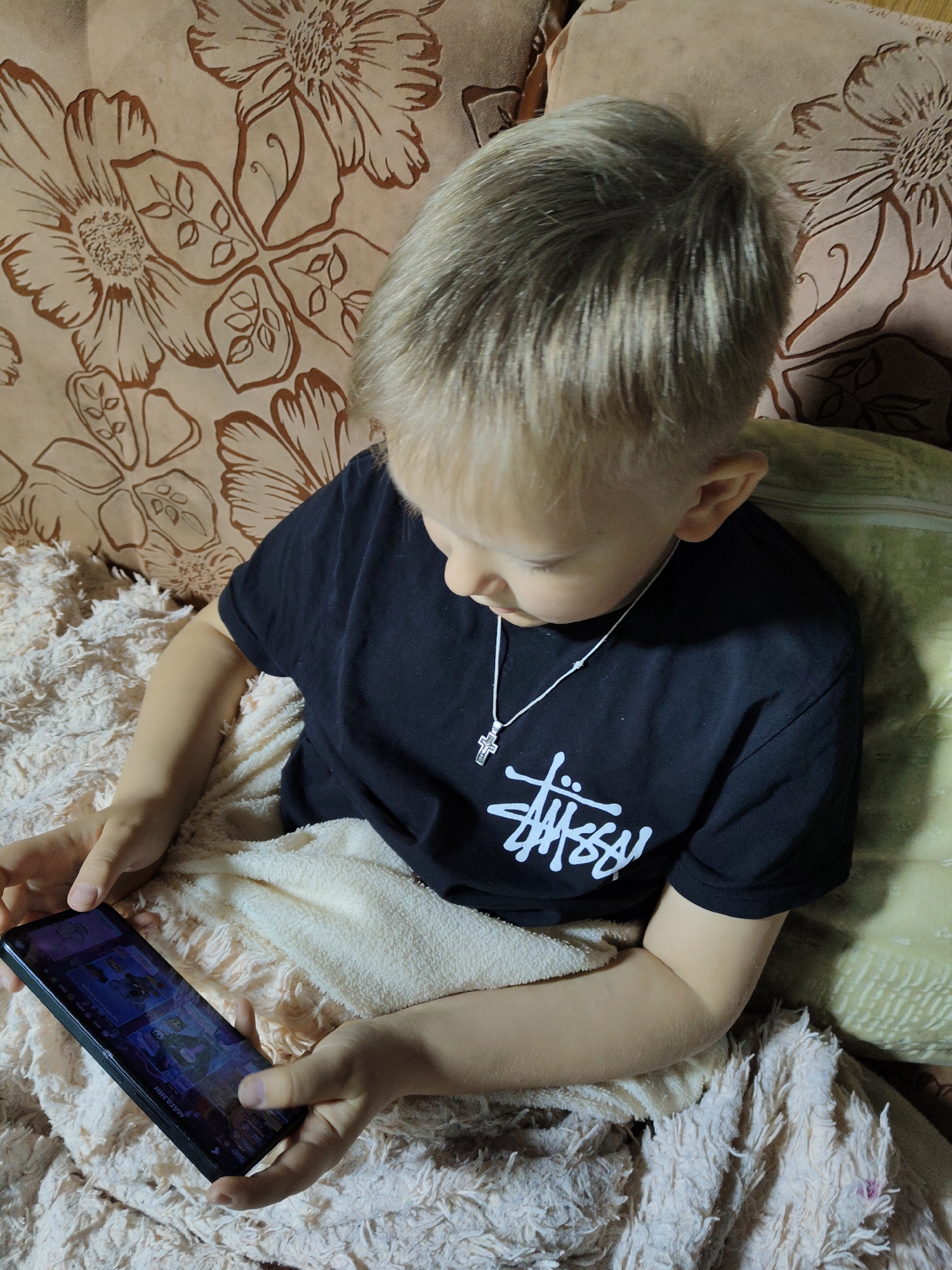 A young child with blond hair is sitting on a patterned couch, covered by a light-colored, fuzzy blanket. The child is wearing a black t-shirt with a white "Stussy" logo and a silver cross necklace. They are holding a black smartphone and looking down at the screen. The couch has a floral pattern in shades of brown and beige.