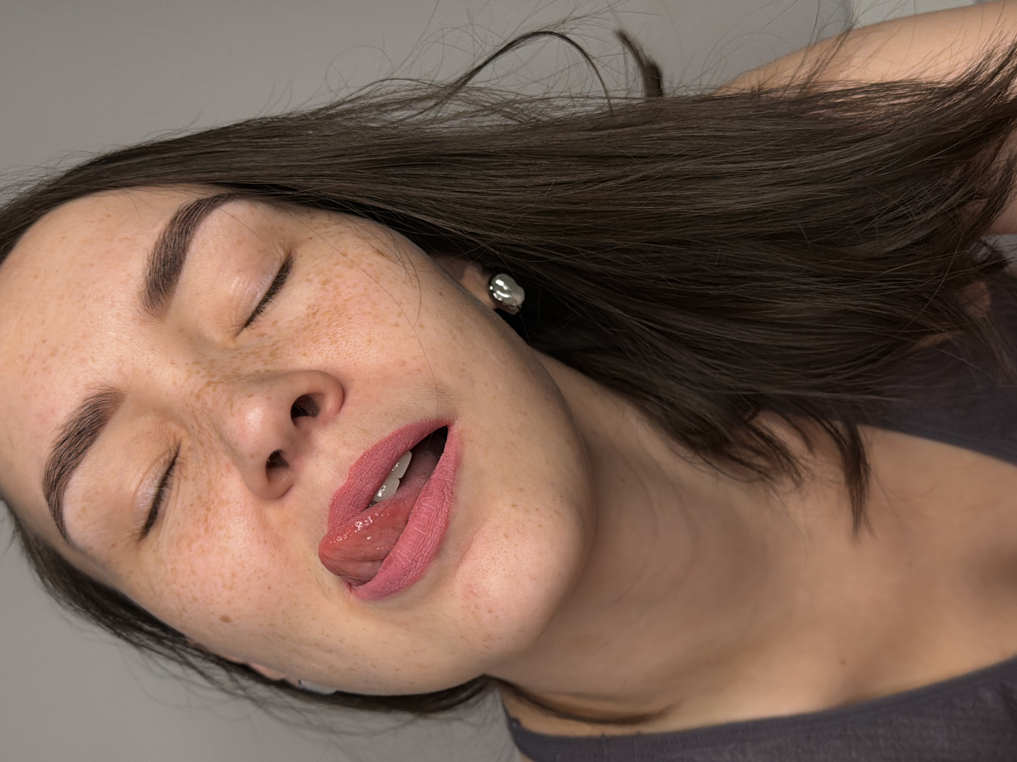 A close-up photograph features a young woman with fair skin and freckles. Her eyes are closed, and her head is tilted. Her tongue is protruding from her mouth, and her lips are coated with pink lipstick. She has dark brown hair and is wearing what appears to be a silver earring. The background is a light grey color. The photograph shows only part of the woman's neck and shoulder, revealing that she is wearing a dark grey top. There is no information available in the image to indicate the location in Chaikovsky, Russia.