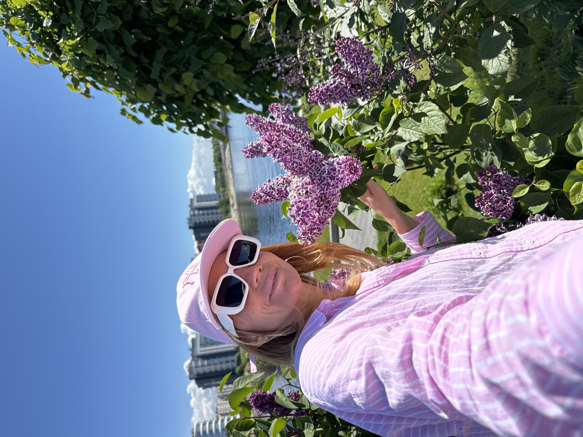 The image shows a selfie of a woman outdoors on a bright, sunny day, likely in Saint Petersburg, Russia. The woman, with light brown hair, wears a pink and white striped long-sleeved shirt, a pink baseball cap with partially visible embroidered text (possibly "NY" or initials), and large white square-framed sunglasses. A golden hoop earring is visible on her left ear.
She is positioned among vibrant purple and white lilac flowers, with her right hand gently near the blossoms, as if admiring them. The background features a wide river, likely the Neva, reflecting the clear blue sky. Modern urban buildings and structures line the distant riverbank under a sky dotted with white clouds. Lush green foliage and grass surround the woman and the lilac bush, indicating a park or riverbank setting. The bright daylight creates strong highlights and shadows, contributing to a cheerful and relaxed atmosphere, suggesting enjoyment of a pleasant spring or early summer day.
