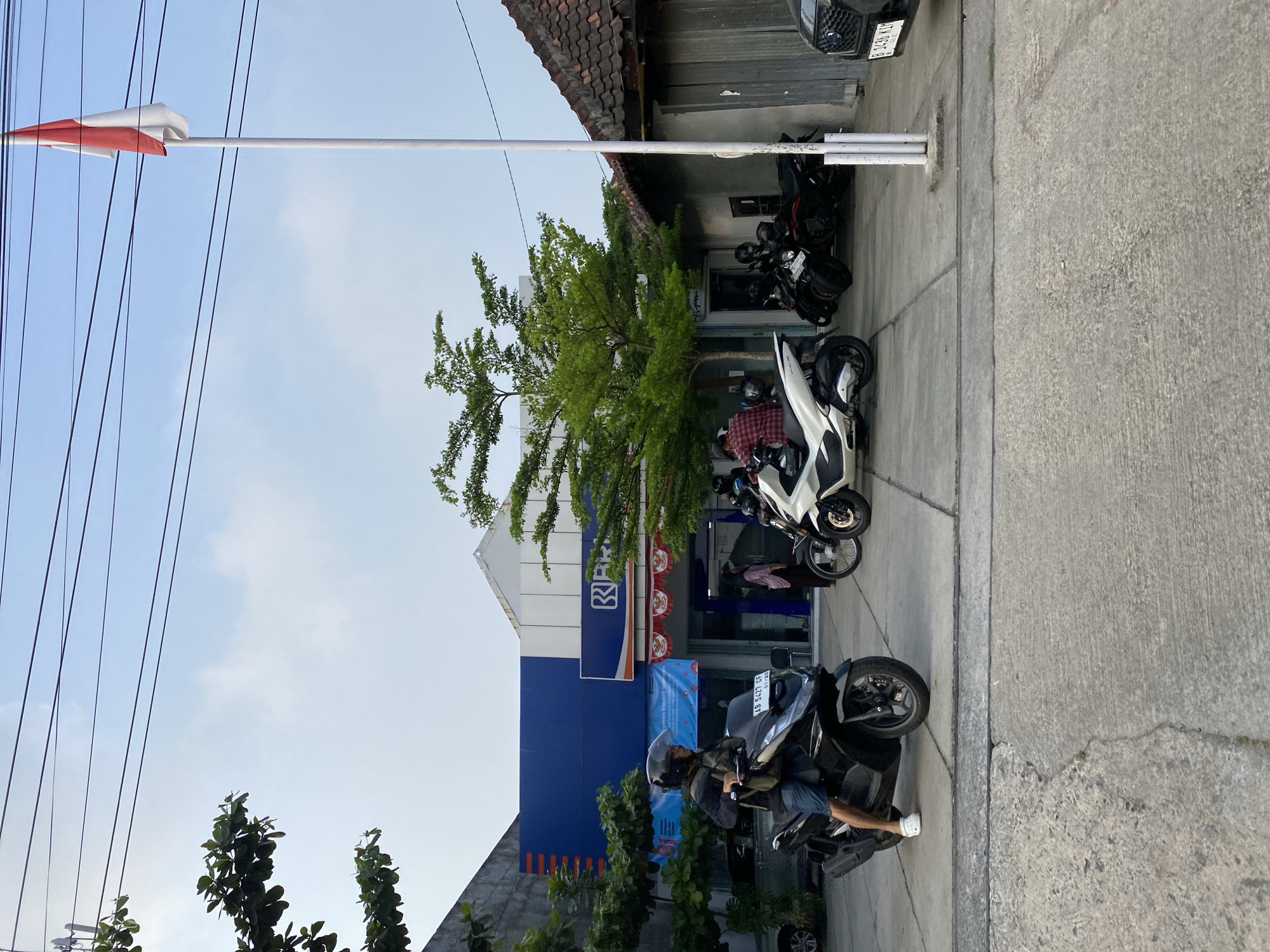 The image depicts an outdoor scene in front of a BRI (Bank Rakyat Indonesia) branch in Kasihan, Indonesia. The bank's facade is blue and white, with the BRI logo visible. Several motorcycles are parked in front of the bank, and people are interacting near the entrance. A man wearing a helmet and shorts is riding a black motorcycle in the foreground. A white motorcycle is parked to the right, and several other motorcycles are visible further back. The Indonesian flag is flying on a flagpole to the right of the bank. The environment appears to be a street or parking area, with concrete ground and some greenery from trees. The sky is overcast.