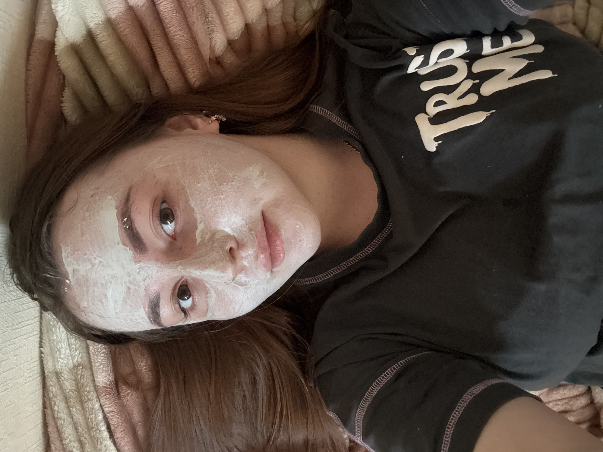 A person is lying down, facing upwards. They have a white facial mask applied. The person is wearing a black t-shirt with the white text "TRUST ME". The person is lying on a textured beige and pink blanket. The background provides no evidence of the city of Izhevsk, Russia.