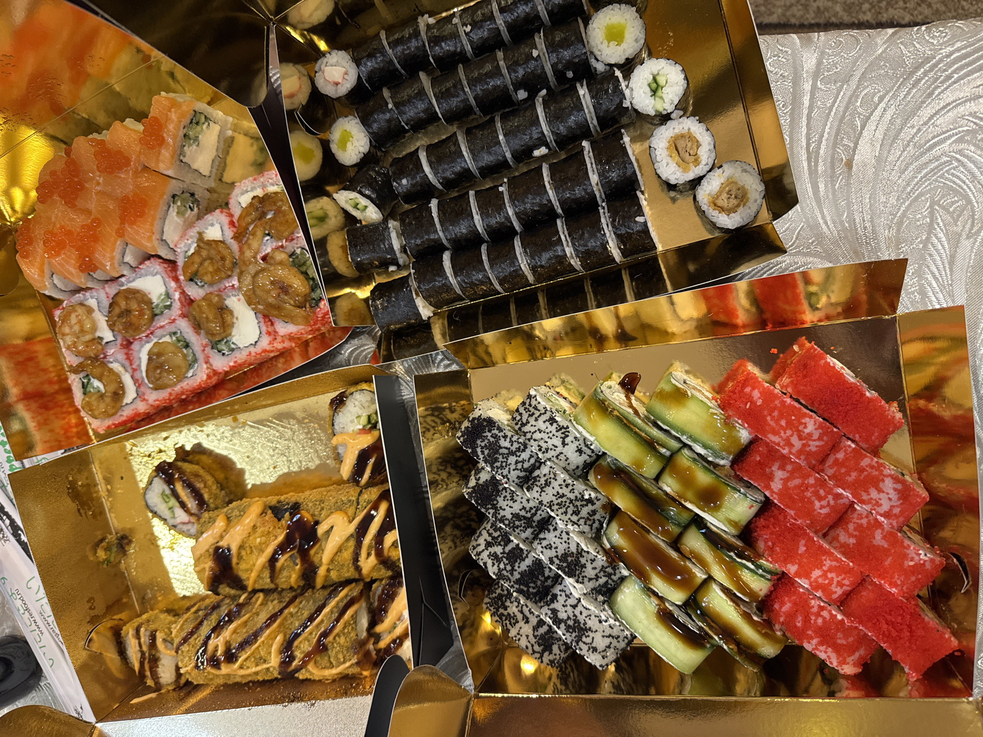 The image shows four golden boxes filled with sushi. The sushi varieties include maki rolls (some covered in seaweed, others in red or black roe), California rolls (with avocado and crab), and fried rolls with a brown sauce drizzle. The scene appears to be indoors, possibly at a residence or restaurant in Yerevan, Armenia, as indicated by the domain name "ru.restoran.www.res..." printed on one of the paper sheets. The table covering underneath the trays is a light, patterned fabric.