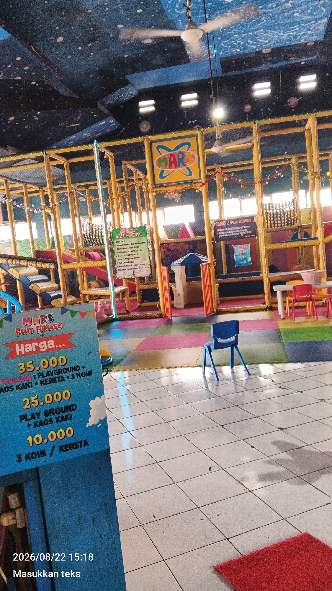 The image shows the interior of a children's indoor playground called "Mars Fun House" located in Malang, Indonesia. The playground features a large, multi-level soft play structure with yellow poles and netting, a blue plastic chair in the foreground, and a red rug. A sign in the foreground displays pricing information for various activities, including "Playground + Socks" for 35,000 IDR and "Playground + Socks" and "Train" for 25,000 IDR. Another sign indicates "3 Coins / Train" for 10,000 IDR. The ceiling is painted with a dark blue starry sky motif and has ceiling fans. In the background, tables and chairs suggest a waiting or seating area. The floor is tiled with a checkered pattern in light and dark squares. A date and time stamp "2026/08/22 15:18" and the text "Masukkan teks" are overlaid on the bottom left of the image.