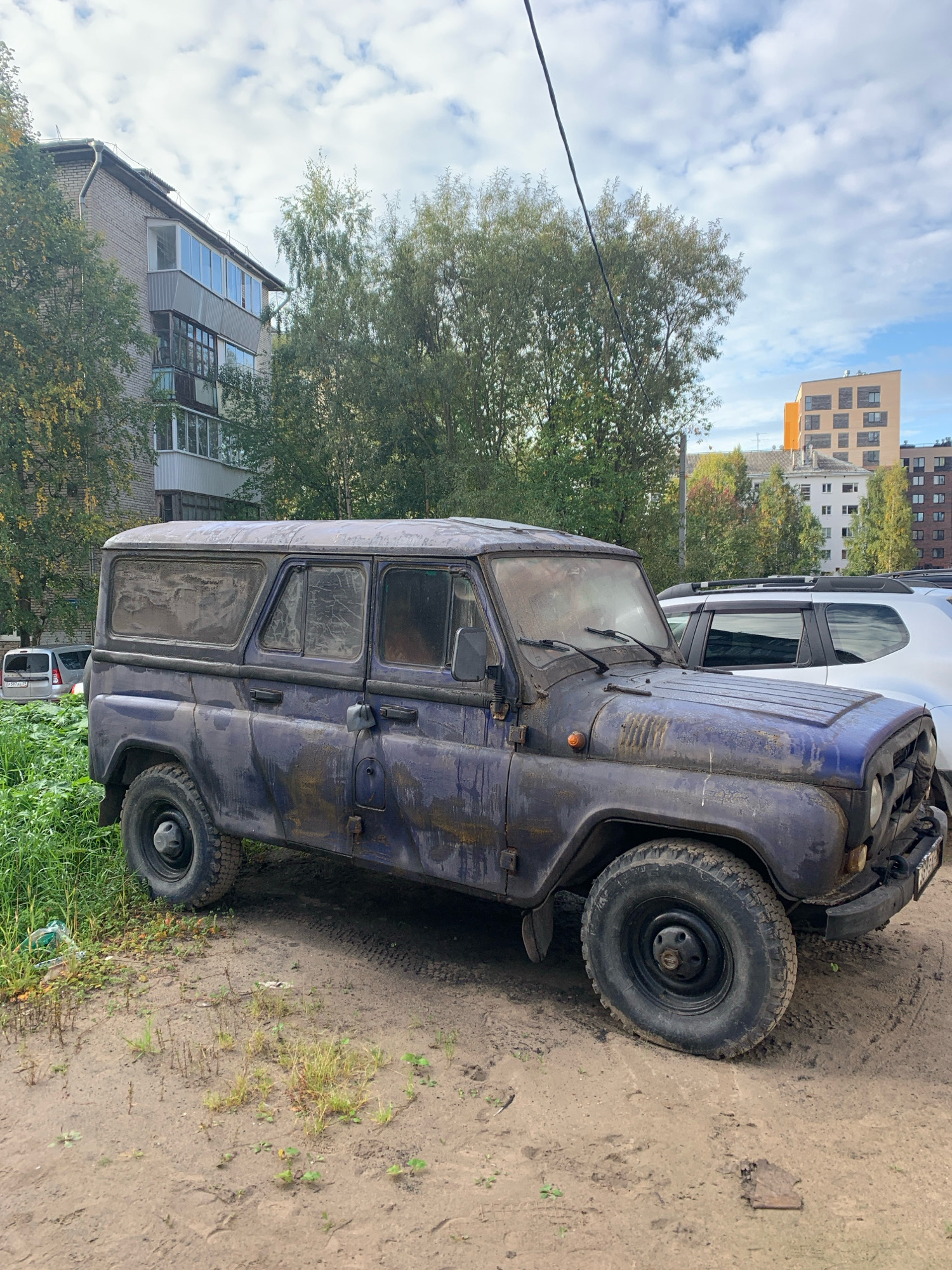 This image captures a blue, weathered UAZ-469 off-road vehicle parked on a dirt lot in Arkhangelsk, Russia. The vehicle exhibits signs of age and disuse, with peeling paint and dirt accumulation. To the left, a multi-story brick apartment building with balconies can be seen, partially obscured by green trees. Further in the background, a modern, multi-story residential building with an orange facade stands against a cloudy sky. Another white vehicle is parked behind the UAZ. The ground is a mixture of dirt, gravel, and sparse vegetation.