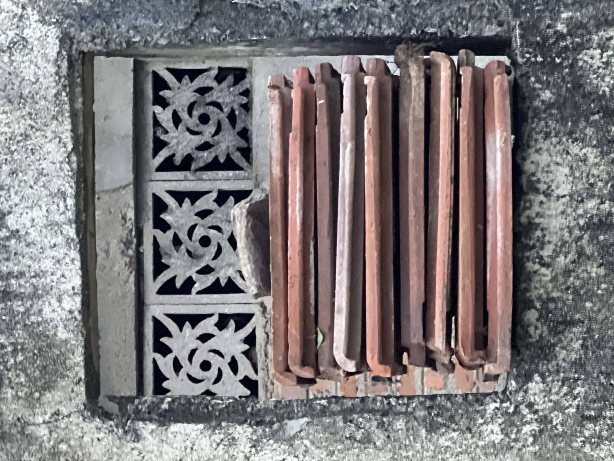 The image shows a close-up, eye-level shot of a portion of a weathered, textured, grey concrete wall. Within the wall, there is an opening covered by a decorative, grey metal grate with three square sections, each featuring an intricate floral or starburst pattern. Below the grate, a stack of approximately twelve reddish-brown, clay roof tiles is piled horizontally, obscuring the lower part of the opening. A small, greenish leaf is visible on one of the tiles. A single grey, rough-surfaced stone rests on top of the grate, slightly off-center to the left. The concrete wall exhibits signs of age and weathering, with visible cracks and a mottled texture. The overall scene appears to be an exterior architectural detail.