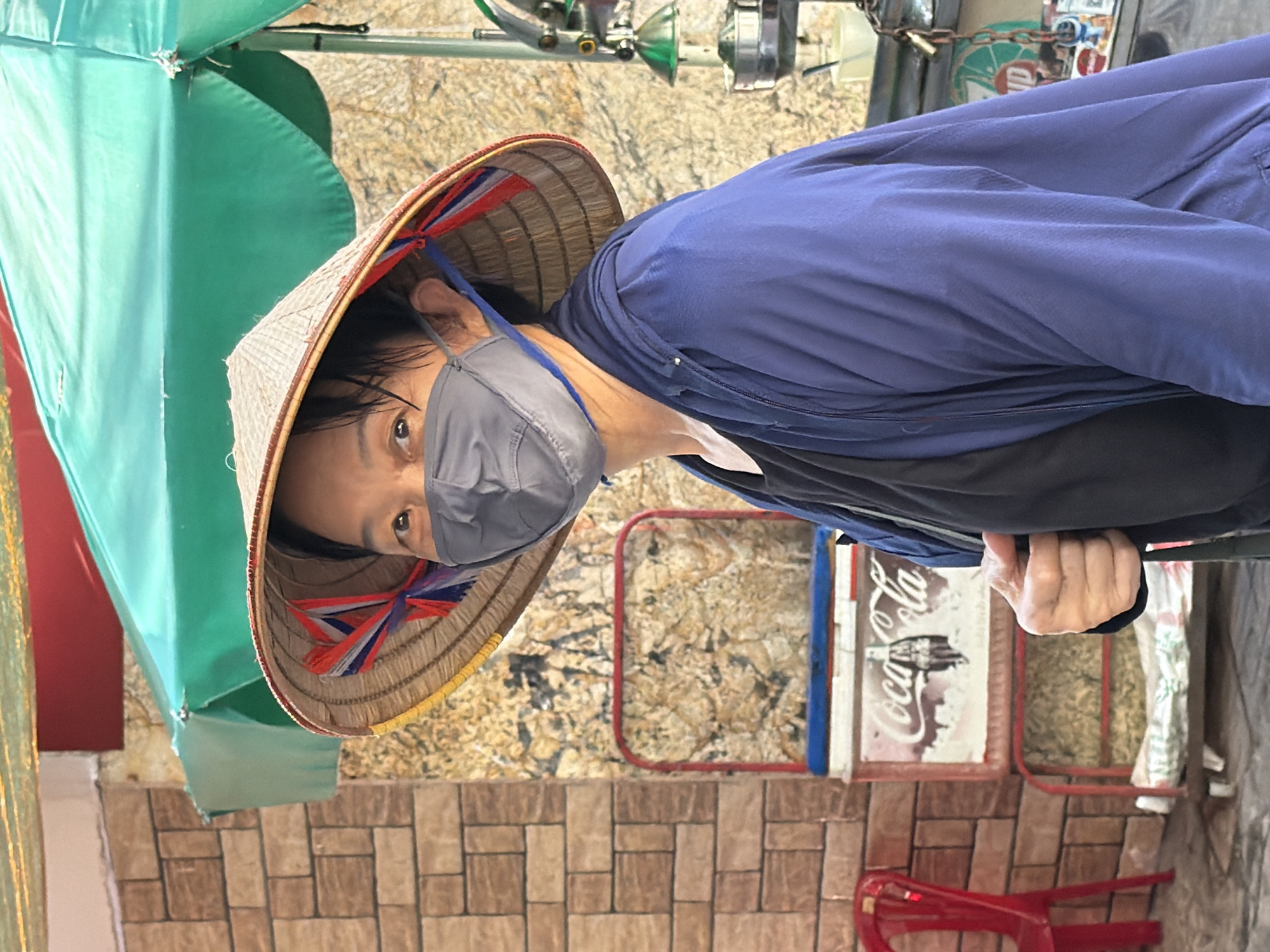 A person wearing a conical hat and a face mask stands in front of a tiled wall and a Coca-Cola cooler cart. The person is wearing a blue jacket over a black shirt. A red chair is visible to the left. A green umbrella shades the scene. The background shows a stall with various items and a lock. The location is identified as Nha Trang, Vietnam.