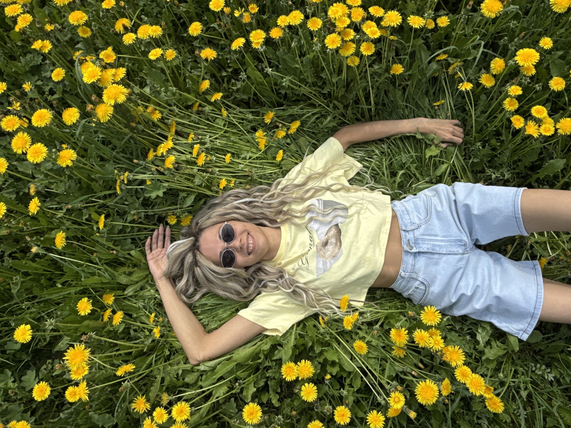 A woman lies in a field of dandelions, dressed in a yellow t-shirt and light-blue shorts. She wears round sunglasses and smiles. The field is filled with blooming dandelions amidst green grass. The location is unknown.