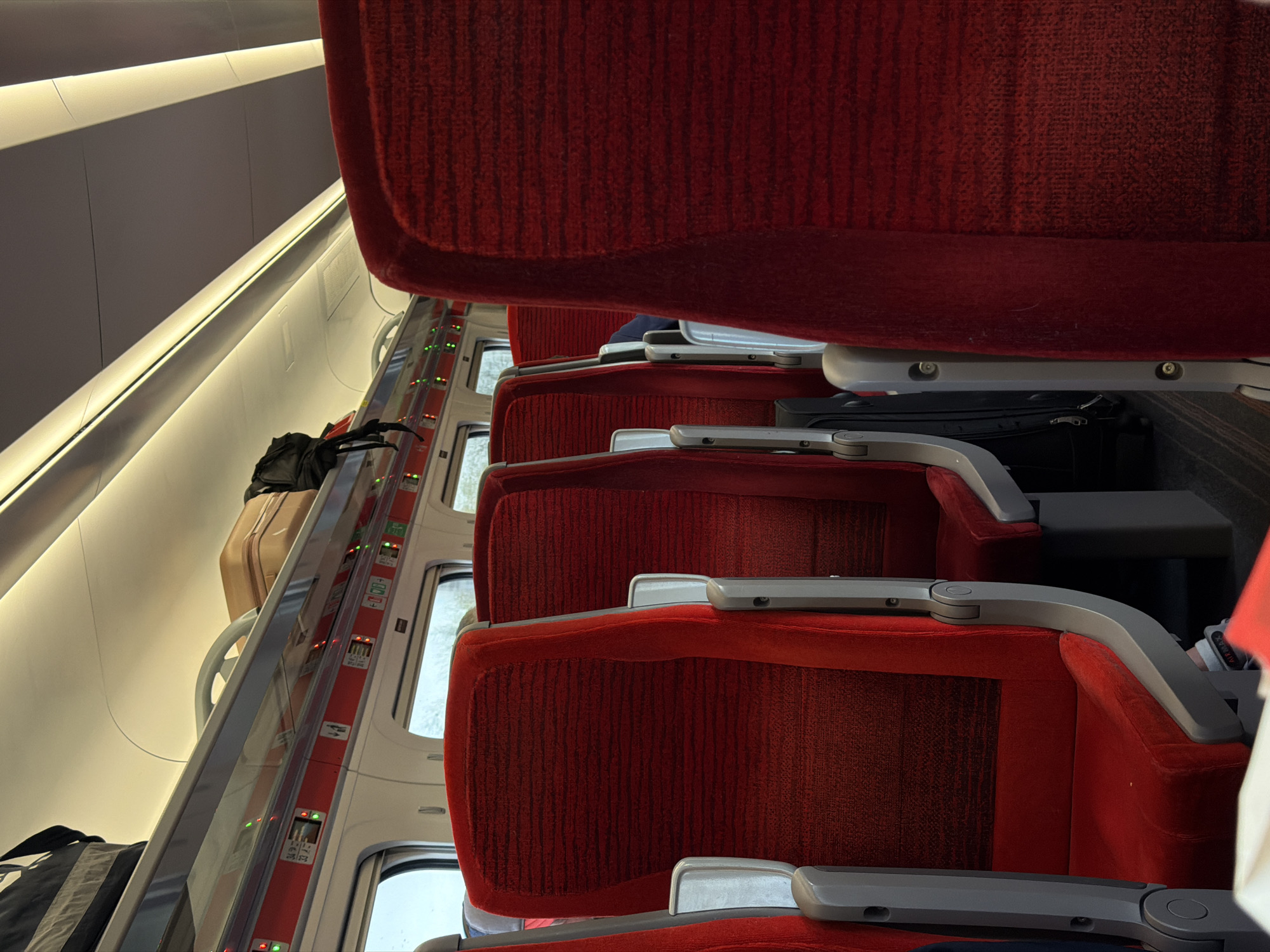 The image shows the interior of a train carriage, likely in Doncaster, United Kingdom given the context. The focus is on a row of red fabric-covered seats with gray armrests. Luggage is visible on the overhead racks, including a black bag and a tan suitcase. The train windows are visible, although the view outside is obscured. Indicator lights above the windows are illuminated.