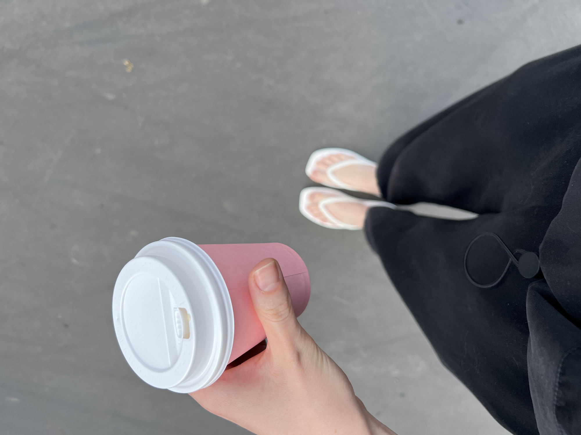 A first-person, high-angle perspective photograph depicts a person holding a pink and white disposable coffee cup with a white lid. The person is wearing black clothing and white flip-flops. The surface underfoot appears to be gray concrete. There is no information to confirm the location in Khimki, Russia.