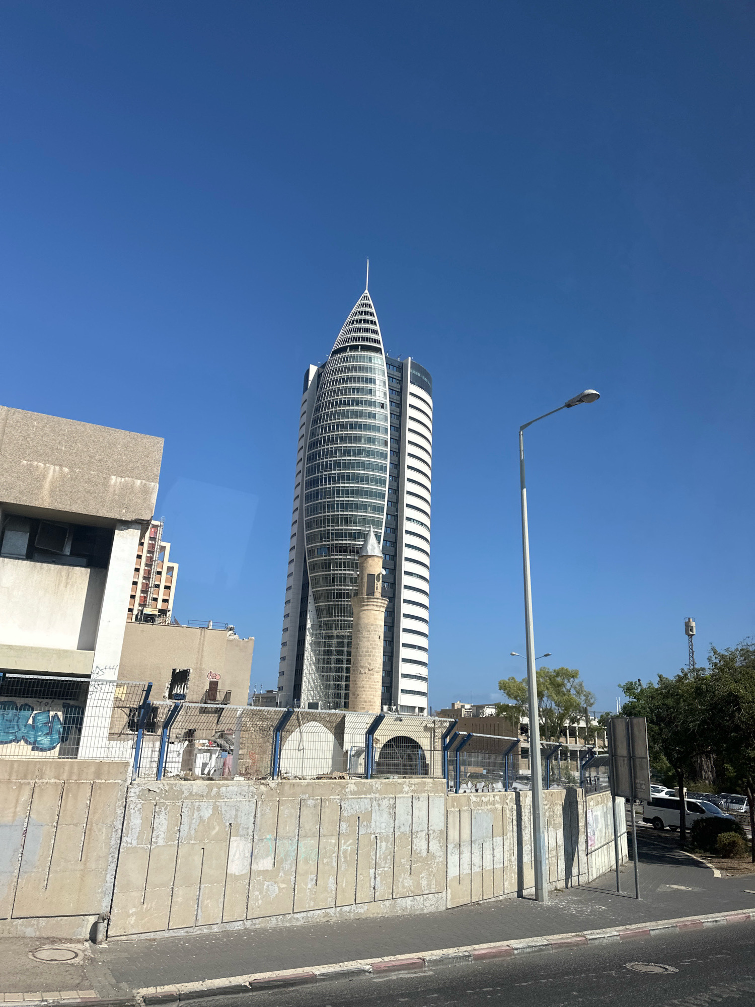 This image depicts a street view in Haifa, Israel, dominated by a modern, tall, curved glass building with a pointed top. In front of this skyscraper, a traditional minaret stands, creating a juxtaposition of old and new architecture. The foreground features a concrete retaining wall with visible signs of wear and graffiti, bordered by a sidewalk and a road. A streetlamp is prominently positioned on the right side of the frame, with trees and other buildings visible in the background under a clear blue sky. There are no people visible in the image.