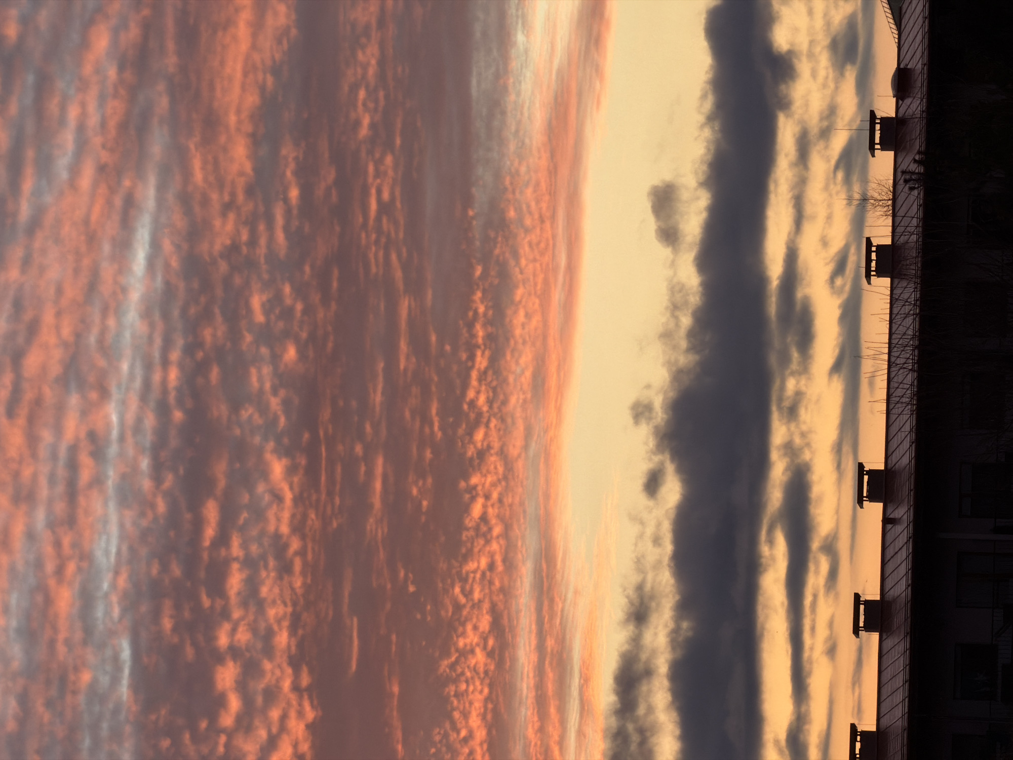 The image depicts a sunset over the Dreiliņi district of Riga, Latvia, with a view of the sky above a building. The sunset is characterized by altocumulus clouds, displaying a range of colors from reddish-orange to grey. The building is silhouetted against the sky, and features elements of urban architecture such as roof-mounted structures, possibly ventilation systems or skylights. The environment is a city with a view of the sky.