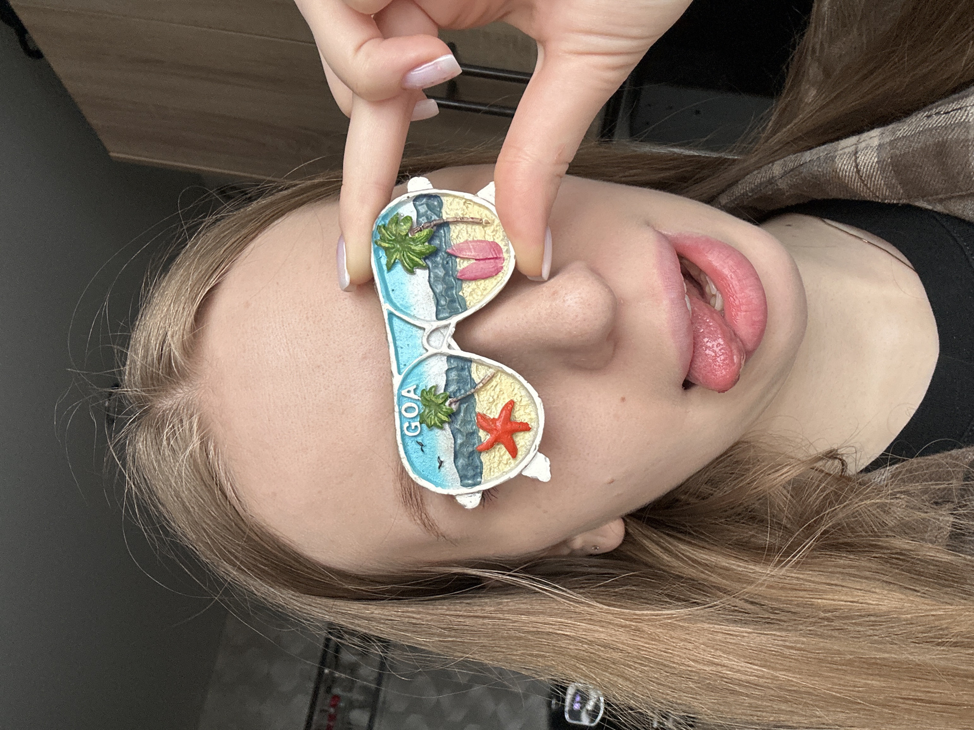 A young woman with blonde hair is lying on her side, sticking her tongue out. A pair of decorative sunglasses with a beach scene design is held over her eyes by a hand with manicured nails. The sunglasses are labeled "GOA." The background appears to be an interior space with a wooden surface. The image does not indicate the location of Chaikovsky, Russia.