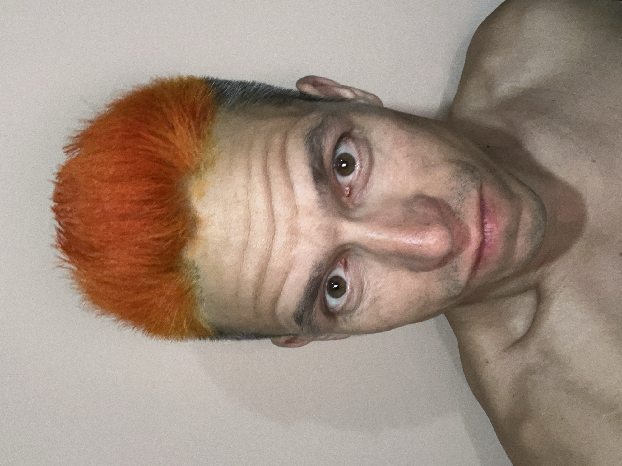 A man with orange hair is angled sideways. The background is plain and light. His body is partially visible. The location is unknown.