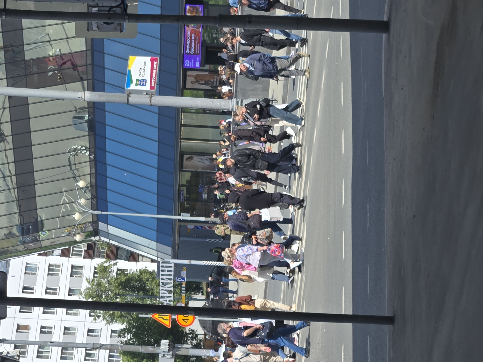 The image shows a street scene in Moscow, Russia, likely near the intersection of Prospekt Mira and Krekshinskaya Street, based on the visible signage. A group of people are crossing a street at a marked pedestrian crossing. They are dressed in casual summer clothing, including t-shirts, jeans, and jackets. Some are carrying bags or purses. The background features a modern building with a glass facade reflecting the sky and surrounding structures. Traffic signs, including a "40" speed limit sign and a warning triangle sign, are visible. A street lamp and traffic lights are also present. The overall atmosphere suggests a busy urban environment on a sunny day.