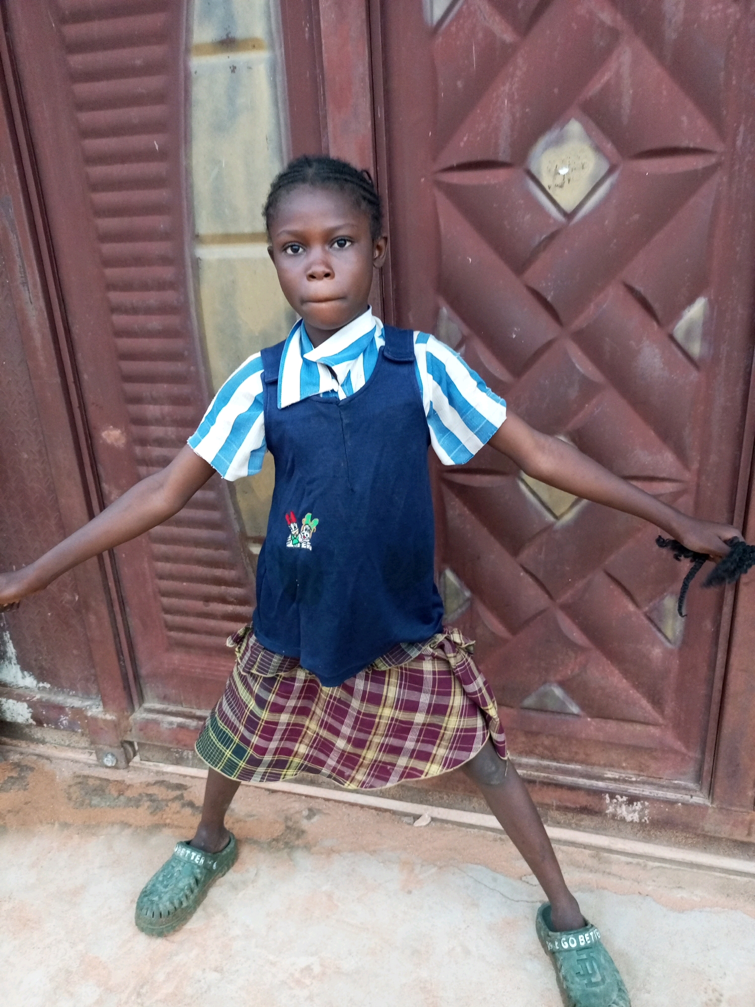 A young girl with braided hair stands with her arms outstretched in front of a dark red, diamond-patterned metal door in what appears to be an outdoor setting, possibly a school or residential area, in Tempure, Nigeria. She is wearing a blue and white striped shirt, a dark blue sleeveless vest with a small colorful emblem, and a plaid skirt in shades of red, yellow, and green. On her feet, she wears dark green clogs with text visible on the left shoe. The ground is a light-colored, uneven surface. The girl's left hand is extended outwards, holding a dark, braided material. Her posture is wide-legged, and she is looking directly at the camera with a neutral expression.