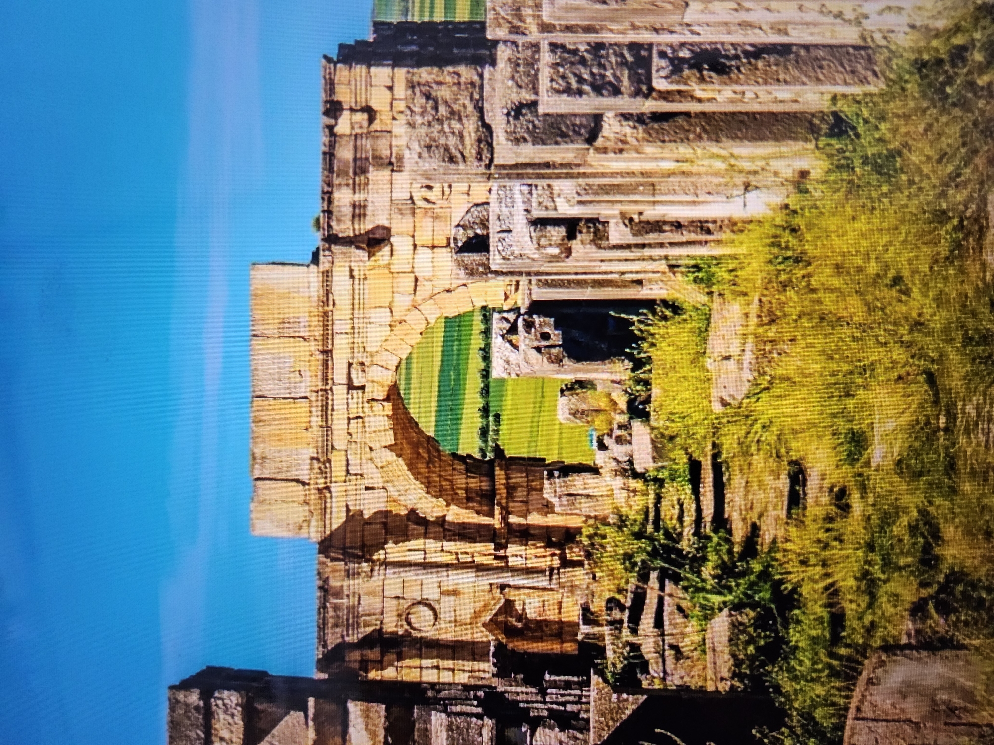 The image shows the ruins of an ancient stone archway against a bright blue sky. The archway is constructed from large, weathered stone blocks, with intricate carvings visible on some of the surfaces. Through the arch, a vibrant green field with rows of vegetation can be seen, suggesting a rural or agricultural landscape beyond the ruins. The foreground is filled with overgrown grass and more scattered stone debris, indicating a site that has been abandoned and reclaimed by nature. The overall impression is one of historical grandeur and decay. This location is not in Payagaboh, Indonesia; it is an archaeological site in a Mediterranean or North African region, likely Timgad, Algeria, a UNESCO World Heritage site featuring Roman ruins.