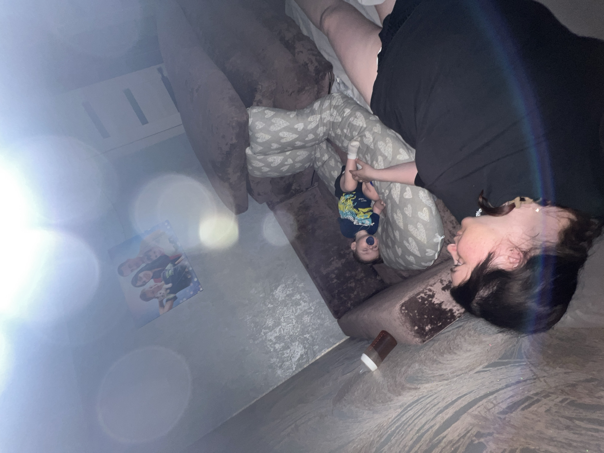 A woman lies on a brown couch with patterned cushions, holding a baby by the legs. The baby, wearing a blue shirt, has a pacifier in its mouth. Next to the couch, on the floor, is a baby bottle and a framed photo of people. The room has a white door with vertical indentations. The photo was likely taken indoors, but I do not have sufficient information to determine that this location is in the city of Noviy, Russia.