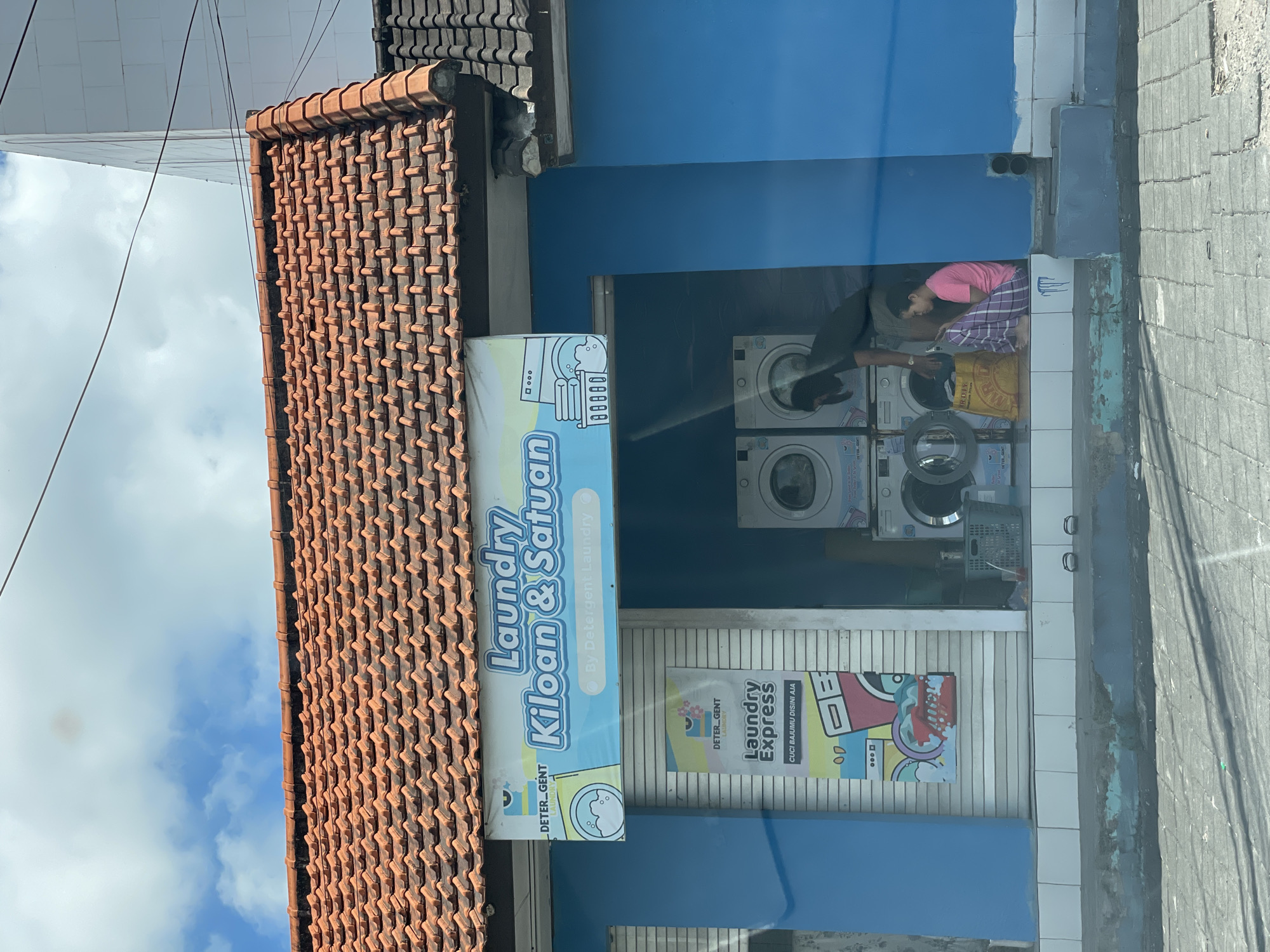 This is a full, eye-level shot of a bright blue laundry establishment located in the city of Kuta Selatan, Indonesia. The establishment has a traditional orange tiled roof with a white and light blue sign that reads "Laundry Kiloan & Satuan" and "By Detergent Laundry". Underneath the main sign, a smaller banner, also blue and white, features the text "Laundry Express" and "CUCI BAJUMU DISINI AJA".
The interior of the laundry is visible through a large open doorway. Several stacked white washing machines are present. In the doorway, a person with dark hair is bending over one of the machines, seemingly attending to it. To the right, a young woman in a pink shirt and checkered pants is sitting on the floor, looking at her phone. A yellow laundry bag sits next to her. The foreground shows a paved sidewalk in front of the establishment, with some debris and peeling paint on the building's foundation. The sky above is partly cloudy with a bright blue sky.