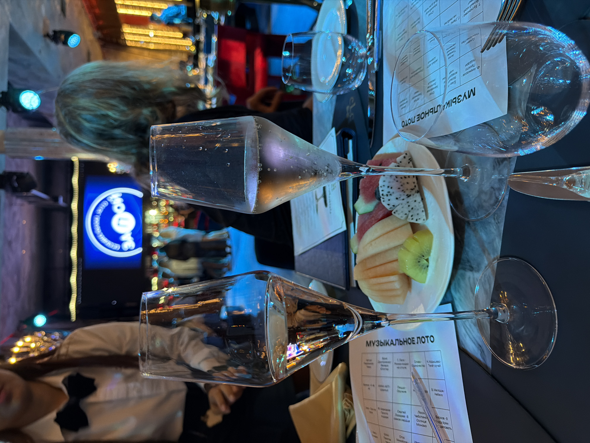 Here is a detailed description of the image:
**Scene:** A table setting at a restaurant, likely indoors. The atmosphere appears to be celebratory and possibly festive, given the presence of sparkling drinks, a fruit plate, and illuminated decorative elements in the background.
**Objects:**
* Several clear wine glasses are positioned on the table, some filled with a sparkling beverage that appears to be champagne or sparkling wine.
* A white plate holds cut fresh fruit, including dragon fruit, kiwi, and melon.
* A menu or game sheet titled "МУЗЫКАЛЬНОЕ ЛОТО" (likely a musical bingo game) lies on the table.
* Cutlery is also present, including a fork and a knife.
**People:**
* A person with blond hair is visible in the upper left corner.
* A person wearing a white shirt and black bow tie is seen in the bottom left.
**Environment:**
* The background is blurred and includes decorative lights, possibly from an outdoor area or a window view. The lighting suggests a nighttime setting.
* There is a blue sign with a logo in the background.
**Details**:
* The image is taken at a close angle to the table setting, focusing on the drinks and food.
* The overall impression is one of an evening dining experience.
* There are no references to Choeng Thale, Thailand in the image.