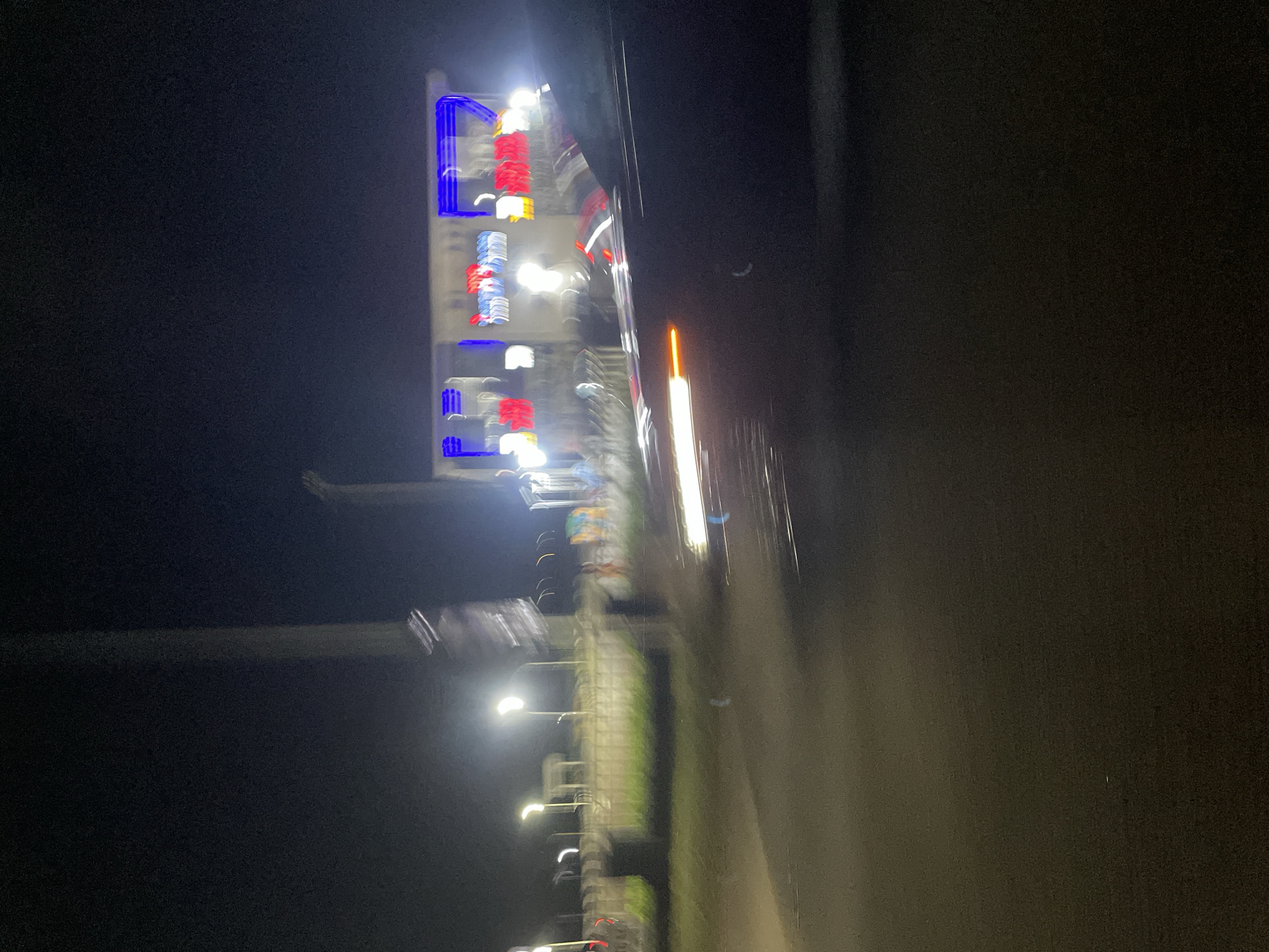 The image captures a highly motion-blurred night scene, likely taken from a moving vehicle or with a moving camera. The perspective is rotated 90 degrees clockwise from its likely original orientation.
The primary subject is a tall, illuminated structure on the left side, featuring prominent bright blue, red, and white rectangular lights arranged vertically. These lights are heavily streaked due to motion blur, making any discernible text or specific symbols unreadable. The structure appears to be a building or a large advertising billboard.
To the right of this structure, a series of streetlights are visible, also appearing as white streaks due to the blur, suggesting a road or elevated pathway runs parallel to the illuminated structure. Below these streaked lights, some blurred green patches indicate vegetation, possibly grass or trees alongside the road. The far right side of the image is largely dark and indistinct, with some vertical light streaks and a dark area that could be a part of the vehicle from which the photograph was captured.
The image conveys the sensation of rapid movement through an urban or semi-urban landscape after dark. The low light and significant motion blur create an abstract, impressionistic view of city lights at night. No people or specific identifying location cues are clearly visible within the frame, beyond the general characteristics of a bustling night environment.