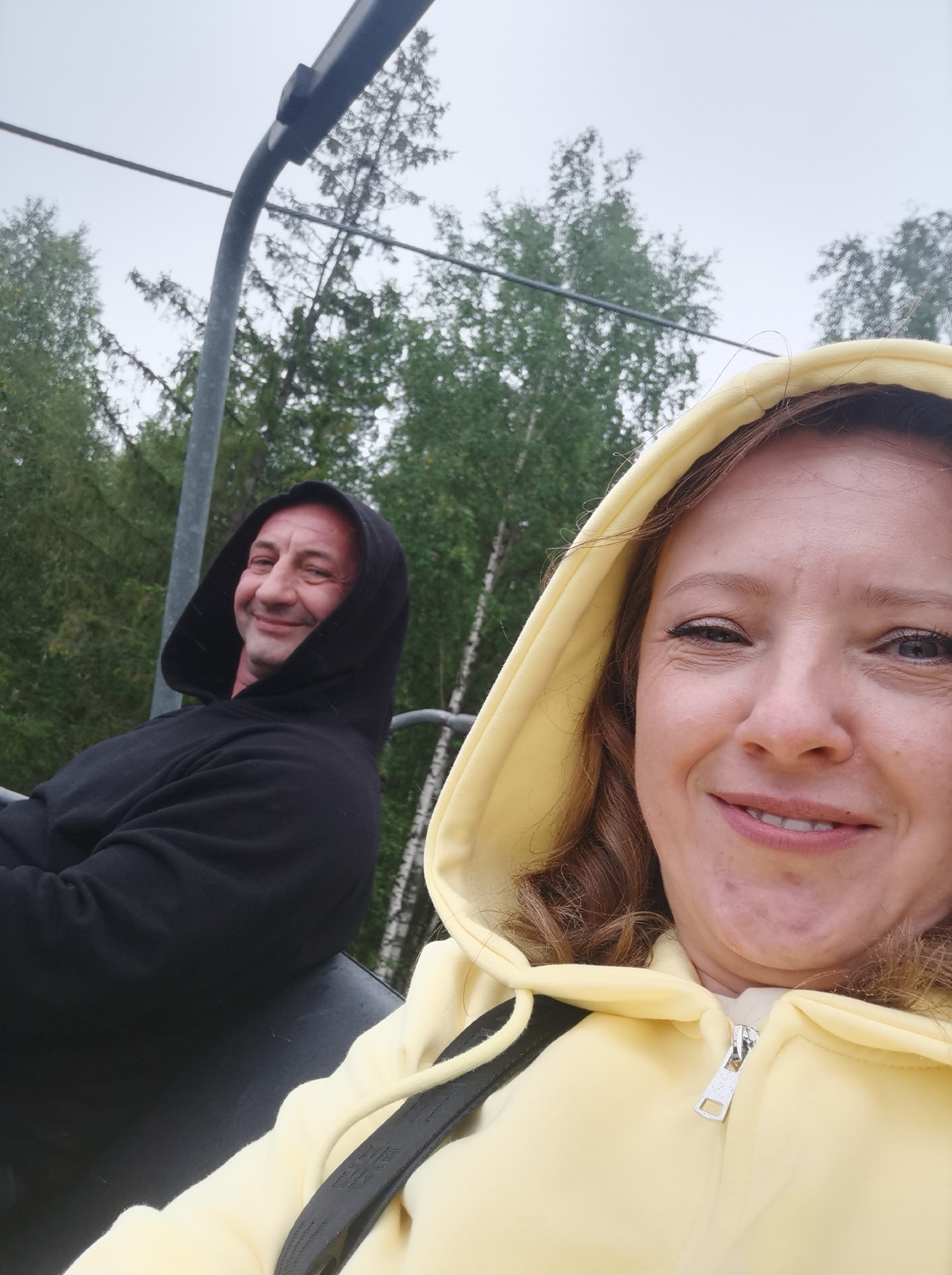 Two individuals, a man and a woman, are captured in a selfie while riding a cable car. The man, on the left, is wearing a black hooded sweatshirt and looking to his left with a slight smile. The woman, in the foreground on the right, is wearing a yellow hooded sweatshirt and smiling directly at the camera. The cable car appears to be moving through a forested area with birch and coniferous trees under an overcast sky. This scene is consistent with the scenery found at the Kuban ski resort in Gubakha, Russia, which features a ski lift that operates in the summer for panoramic views of the surrounding mountains and forests.