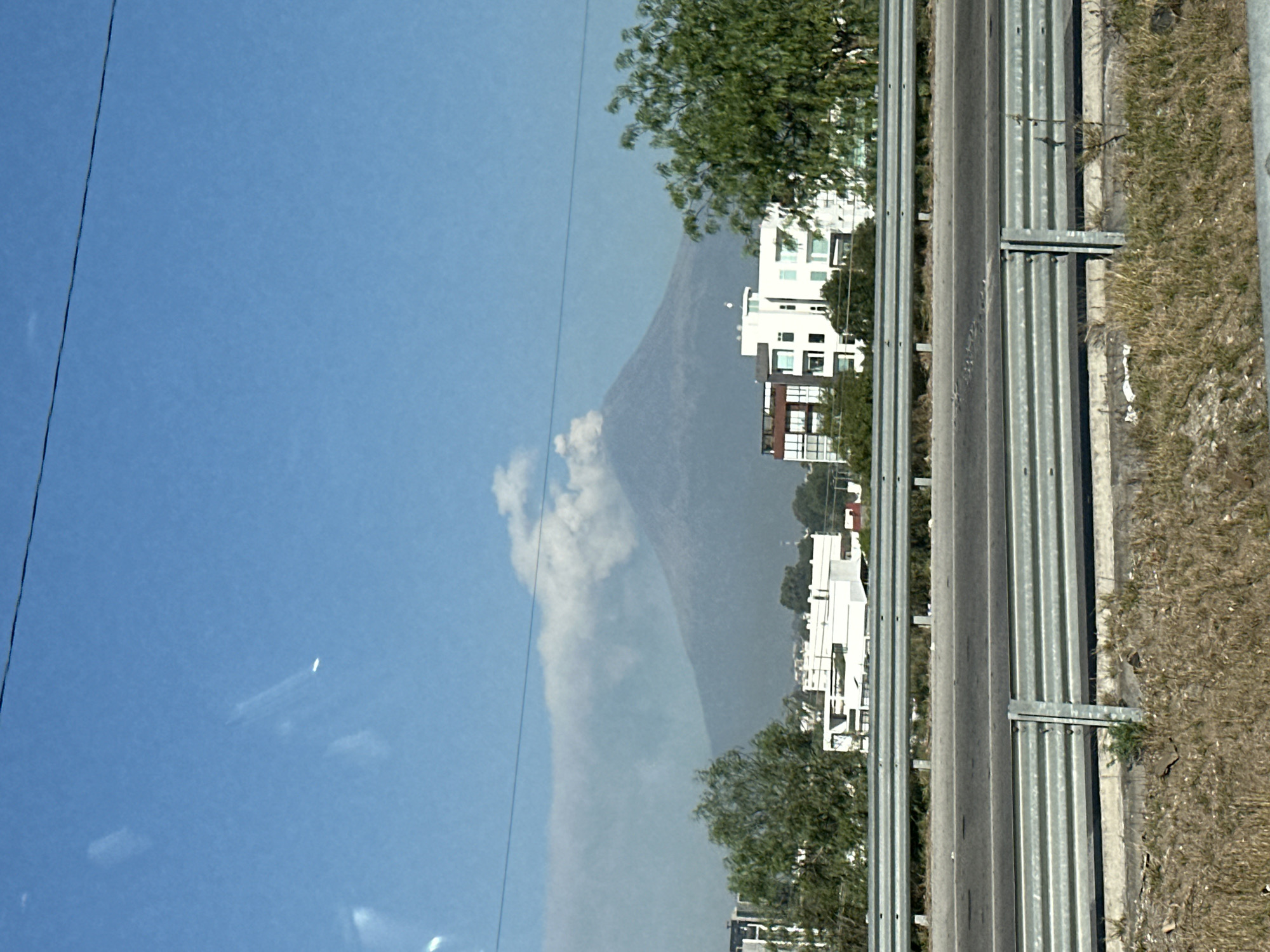 The image captures a daytime scene, likely from a vehicle, looking out at a highway with a prominent mountain or volcano in the background. The location is San Bernardino Tlaxcalancingo, Mexico.
In the foreground, a multi-lane asphalt road runs horizontally across the frame, bordered on its right by a long, corrugated metal guardrail. Adjacent to the guardrail is a strip of dry, brown grass and dirt, indicating a roadside shoulder.
Beyond the road and guardrail, several modern, multi-story buildings, mostly white with some darker sections, are visible, interspersed among green trees. These structures appear to be part of a residential or commercial development.
The dominant feature in the midground and background is a large, hazy grey mountain or volcano, its summit partially obscured by a distinctive white cloud or plume of steam/ash. The sky above is a clear blue with a few scattered white clouds. A thin, dark utility wire or power line stretches diagonally across the upper left portion of the sky.
The bright, natural lighting suggests it is daytime, likely late morning or early afternoon. The scene combines elements of infrastructure and urban development with a striking natural landscape, hinting at the volcanic activity common in the region. No specific human activity is depicted, and there is no visible text.