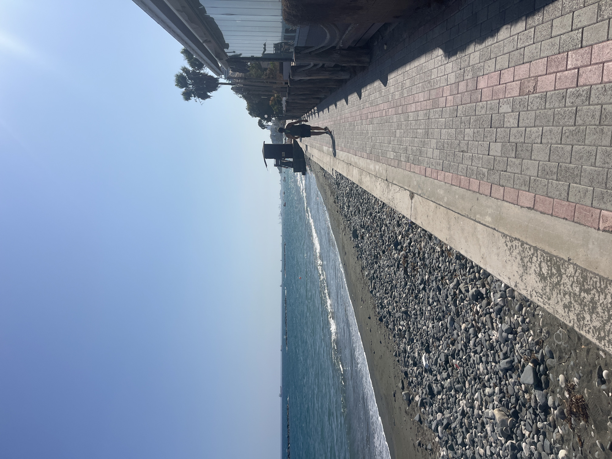 The image depicts a seaside promenade in Agios Tychona Community, Cyprus, under a clear, bright blue sky. The promenade is paved with rectangular bricks in a pattern of grey and pink. To the left of the promenade is a pebble beach leading to the turquoise waters of the sea, where gentle waves are breaking on the shore. In the distance, several cargo ships are visible on the horizon.
On the right side of the promenade, there is a building with large glass windows, suggesting it might be a restaurant or cafe, with a partial view of its exterior seating area. A wooden fence with thick ropes runs along the edge of the promenade, separating it from the building. Palm trees are scattered along the coastline, adding to the tropical feel.
A lone individual is walking away from the camera along the promenade, wearing a dark tank top and shorts, and a bandana around their head. They appear to be casually strolling. Near the beach, there is a small, dark-colored structure that resembles a lifeguard station or a small booth. The overall scene is one of a peaceful day at the beach, with a person enjoying a walk along the coast.