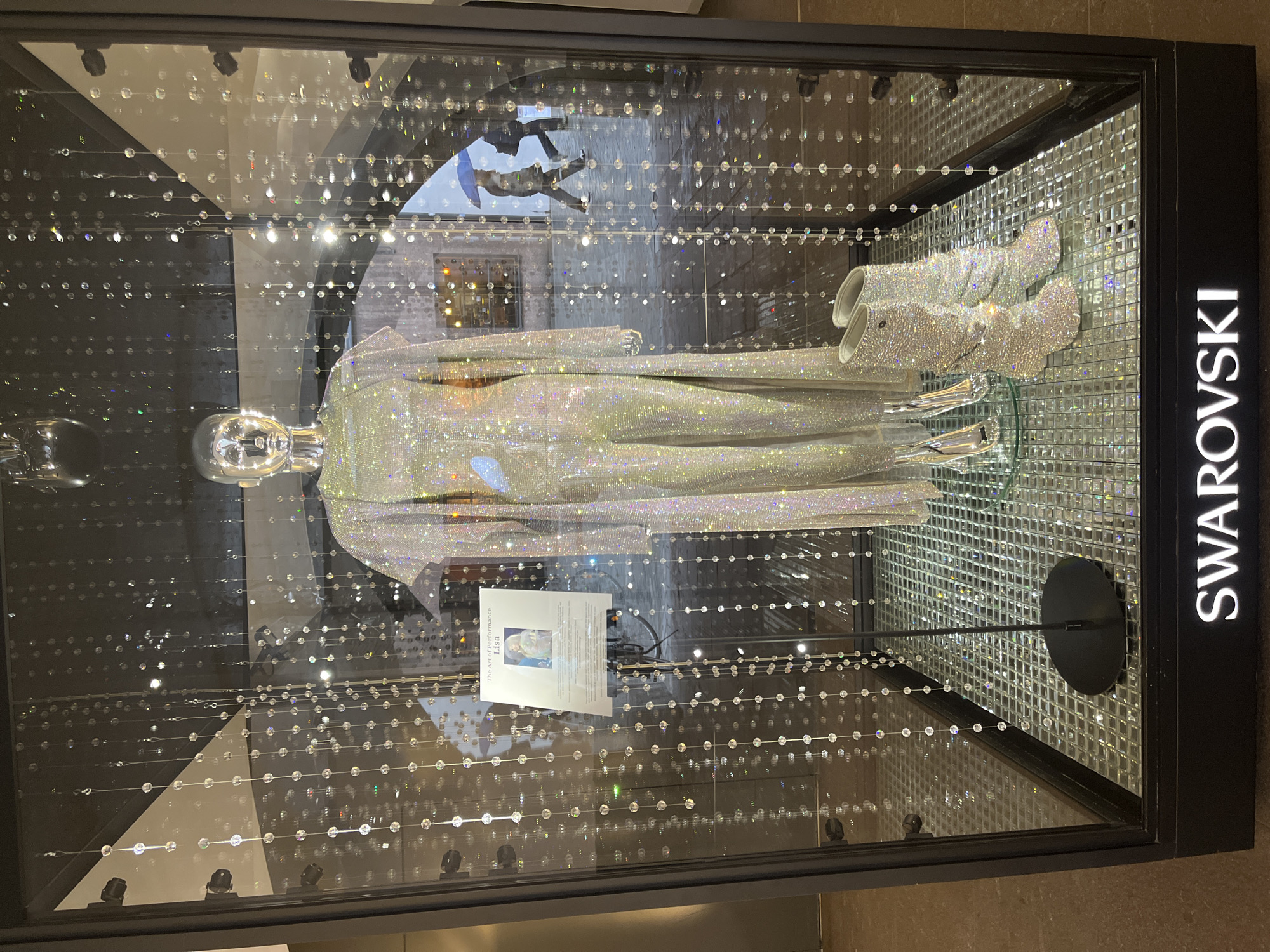A Swarovski crystal-encrusted display case in Innsbruck, Austria, features a silver mannequin adorned in a matching shimmering jumpsuit and a long, flowing robe. Crystal bead strands hang vertically throughout the display, creating a glittering effect. To the left of the mannequin, a small information card with text and an image titled "The Art of Performance Lisa" is displayed. The mannequin stands on a tiled, reflective platform. On the right, a pair of dazzling crystal boots are showcased. The background of the display shows a blurry reflection of people walking outside with umbrellas. The bottom of the display case prominently features the "SWAROVSKI" logo.