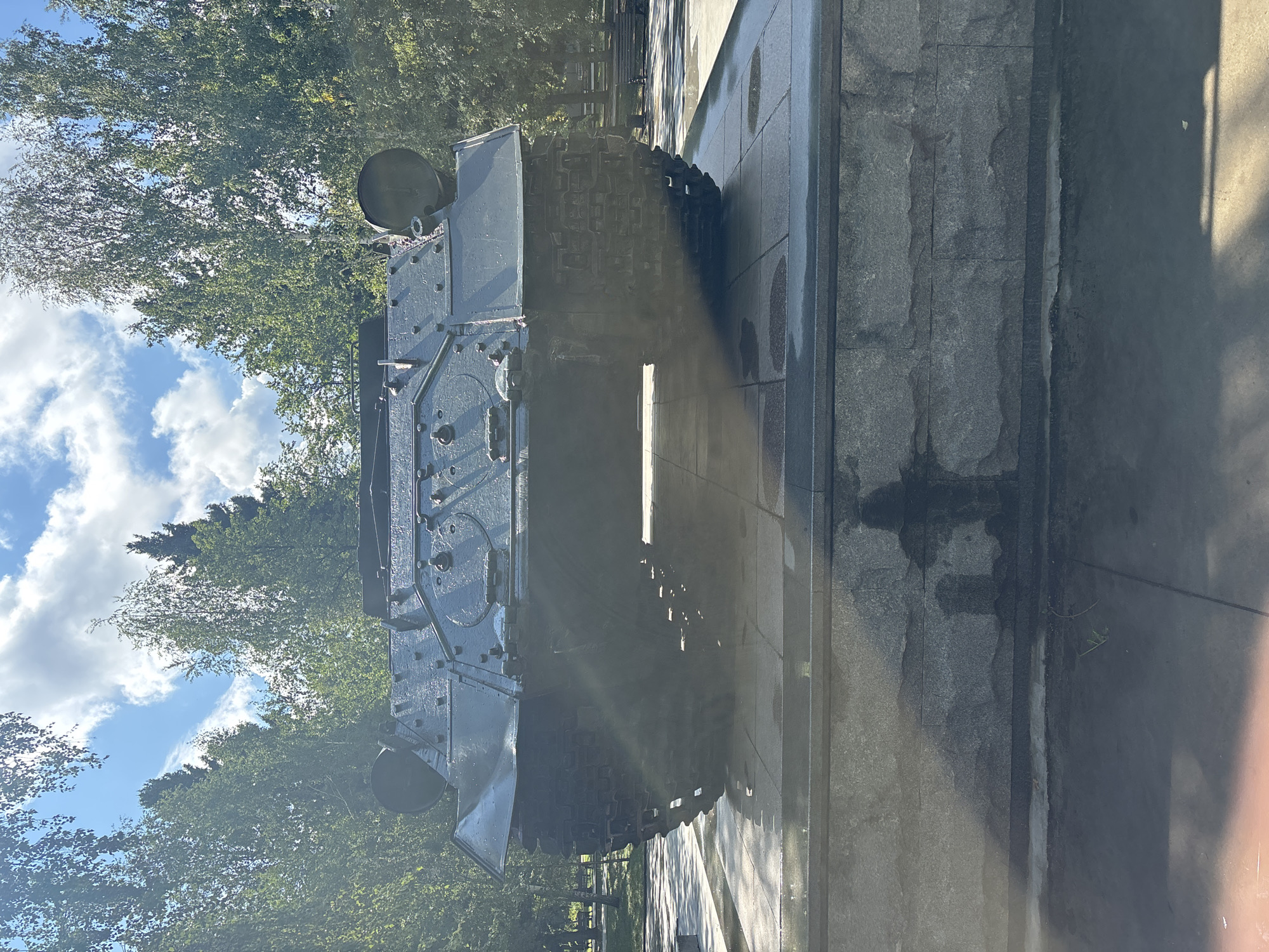 The image shows a monument of a tank in a park in Novosibirsk, Russia. The tank is dark green and appears to be a Soviet-era model. It is displayed on a granite pedestal, which is set on a paved area. The tank is oriented facing the camera, with its tracks prominently visible. The background consists of lush green trees and a blue sky with white clouds. Sunlight filters through the trees, casting dappled shadows on the ground and the tank. In the distance, park benches and more trees are visible, suggesting a public park setting. The scene is peaceful and static, with no people or animals present. The overall impression is of a historical monument in a well-maintained park.