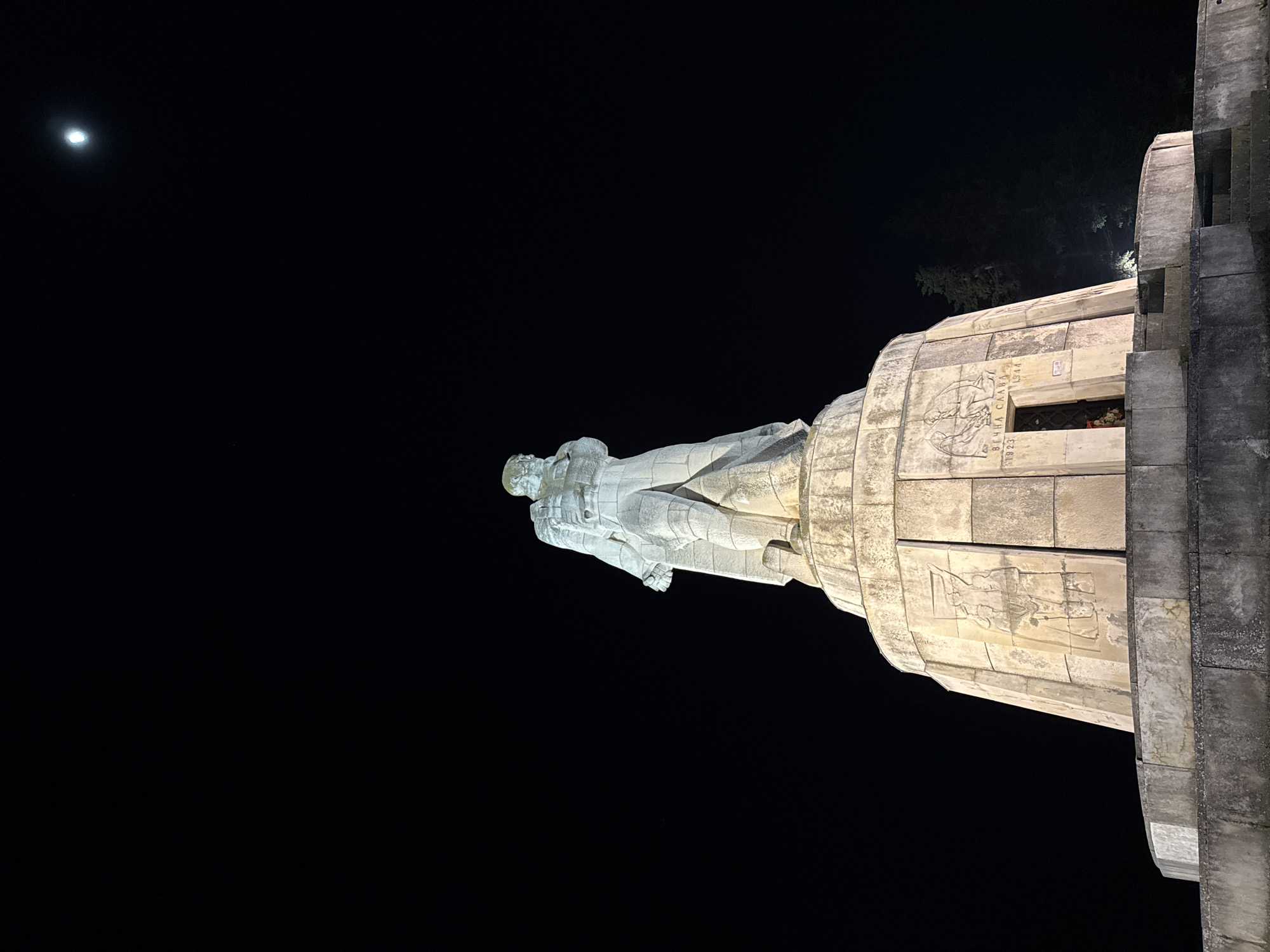 The image displays a prominent stone monument captured at night, with artificial lighting illuminating its features against a deep black sky. The central element is a large, light-colored statue of a standing figure, likely a soldier or hero, clad in what appears to be military attire. This statue is an integral part of a massive, multi-tiered stone structure.
The monument itself is constructed from large, light-toned stone blocks, featuring sculpted relief panels. On one section, visible to the right of the statue, intricate carvings depict figures, possibly soldiers in battle or symbolic scenes. Below these reliefs, Cyrillic text is inscribed, reading "ВЕЧНА СЛАВА," which translates to "ETERNAL GLORY." Adjacent to this inscription are the dates "1923" and "1944." A small, recessed opening beneath the text contains what appear to be flowers, suggesting a place of remembrance or offering.
The time of day is clearly night, evidenced by the dark sky. A bright, distinct moon is visible in the upper left quadrant of the frame, adding to the nocturnal atmosphere. The artificial illumination dramatically highlights the monument's details and textures, creating a strong contrast with the surrounding darkness. Faint outlines of trees can be seen in the background behind the upper sections of the monument, indicating an outdoor setting, possibly a park or public square. The overall impression is one of solemnity and commemoration. The monument is located in Varna, Bulgaria.