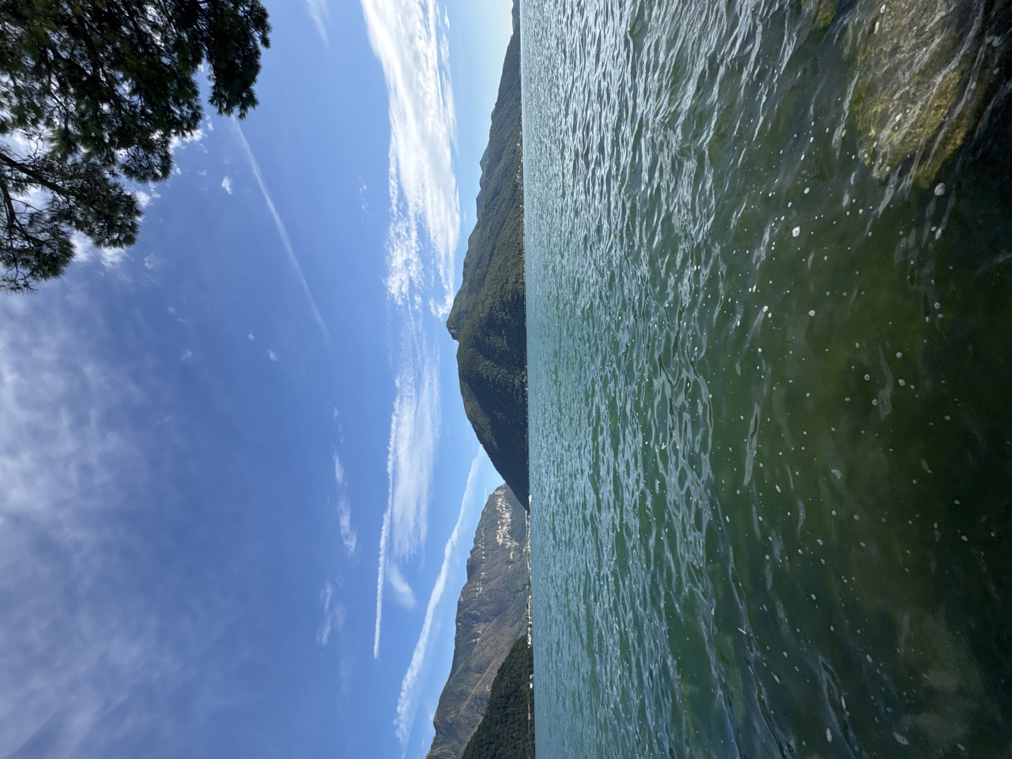 A body of water, likely a lake, is shown in the foreground, with a few rocks visible on the right side near the shore. The water is a deep green color and appears slightly choppy. In the background, a range of mountains covered in green trees rises from the far side of the water. The sky is a clear blue with a few wispy white clouds scattered across it. The image appears to have been taken from a low vantage point, looking across the water towards the mountains. This view is consistent with locations along the shore of Lake Lugano in Melide, Switzerland, specifically looking towards the mountainous landscape across the lake.