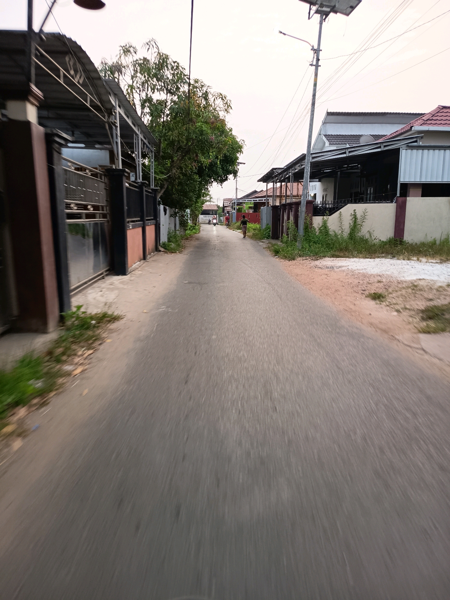 This is a medium-shot, eye-level, outdoor photograph of a residential street in Banjarmasin, Indonesia. The image is taken at dusk, as indicated by the soft, diffused light. The street is paved with asphalt and is slightly blurred, suggesting movement. On the left side of the street, there are houses with dark metal gates and covered carports. On the right side, more houses are visible, some with red tile roofs and others with gray metal roofs. A prominent feature on the right is a tall utility pole with a solar panel and a street light.
In the distance, two people are walking down the street. One individual appears to be a child or a smaller adult, wearing dark clothing and walking ahead. Behind them, a person in a red shirt is walking. A scooter or motorcycle is also visible in the distance, stopped or moving slowly. The environment is characterized by modest residential buildings, scattered vegetation, and a general sense of quiet suburban life. The sky is overcast and pale.