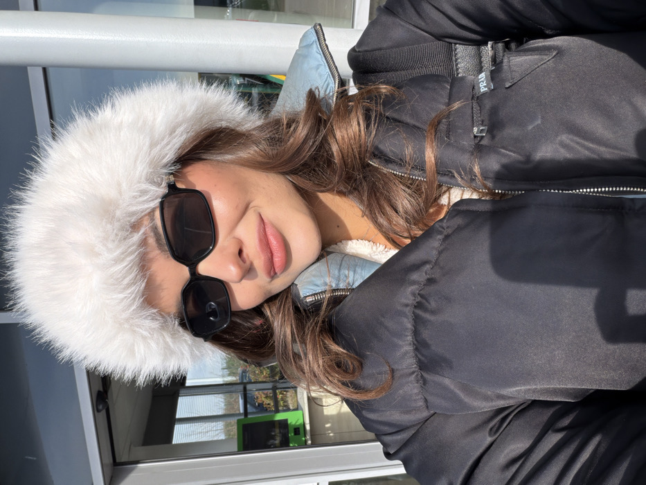A female individual is depicted in a close-up, likely selfie-style, outdoor photograph. She is wearing a prominent white, fluffy faux fur hat that covers most of her head, dark rectangular sunglasses, and a black puffer jacket with a visible silver zipper. Beneath the puffer jacket, a light blue collar and a cream-colored, textured lining are discernible. A black strap from a bag or backpack is visible over her right shoulder, with a black triangular patch on the jacket bearing the "PRADA" logo. The individual has medium-brown, wavy hair and a subtle, closed-lip smile. Strong sunlight illuminates the scene, casting shadows. In the background, visible through large glass windows, are architectural elements characteristic of a modern facility, including white support columns and a bright green electronic kiosk, possibly a parking payment machine. The probable location, given the city of Colombier-Saugnieu, France, and the visual cues, is Lyon-Saint Exupéry Airport.