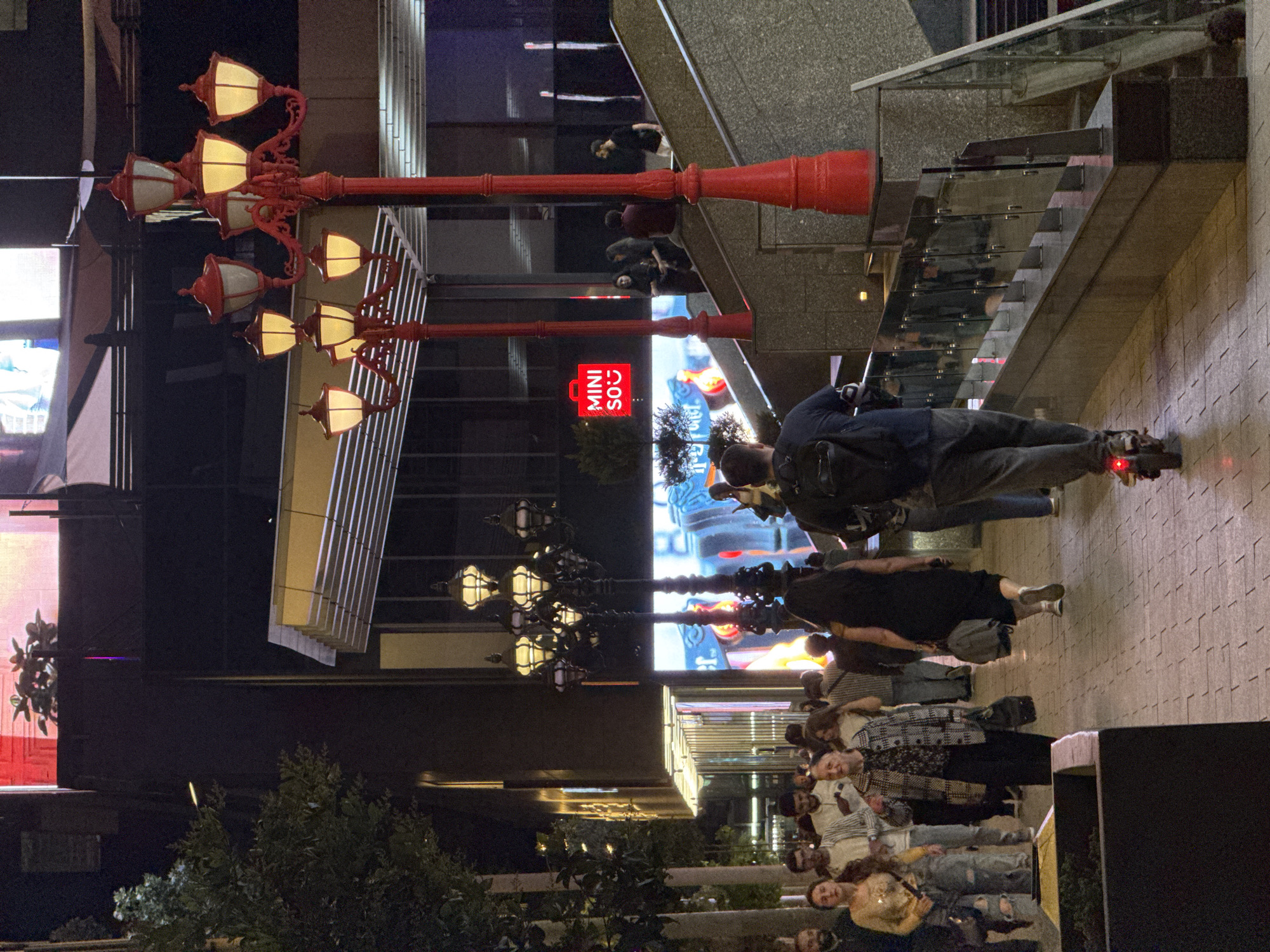 The image depicts a bustling street scene at night in Yerevan, Armenia. A group of people are walking along a bricked sidewalk. One person is roller skating. A row of vintage-style red lampposts casts light on the scene. A shop labeled "MINISO" is visible. Modern buildings and stairways with glass railings form the backdrop.