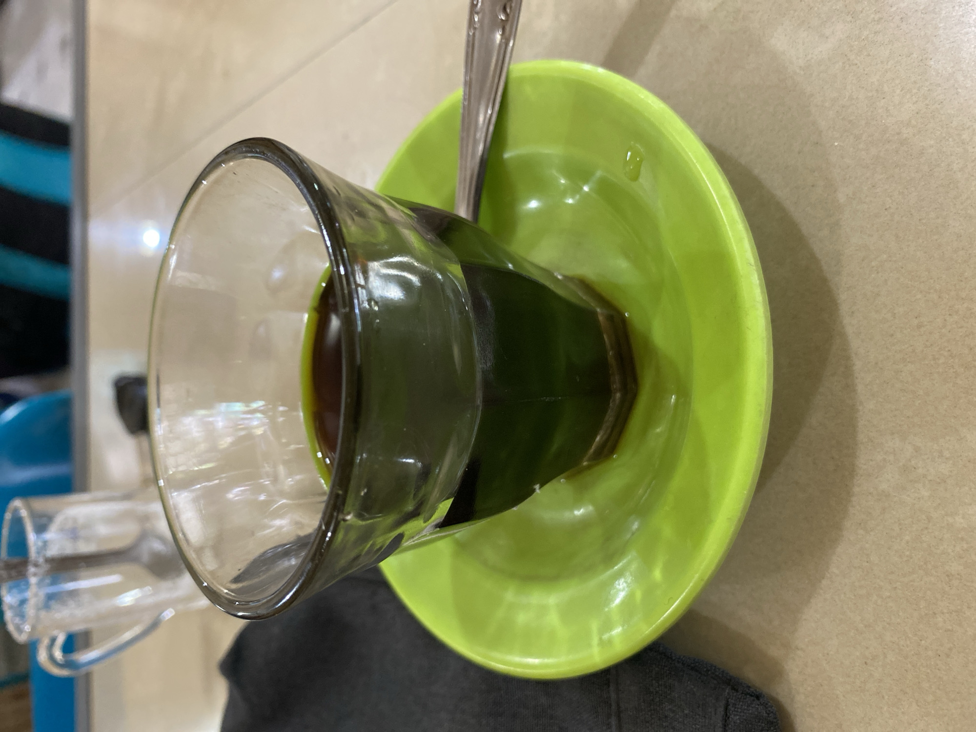 A close-up, eye-level shot shows a small glass filled with a dark liquid, likely coffee, resting on a vibrant green saucer. The glass has a unique hexagonal base and is partially filled, with the liquid reflecting light. A silver spoon rests on the saucer, partially obscured by the glass. To the left of the glass, a dark grey napkin is visible. In the background, out of focus, another clear glass mug with a spoon inside, and some colorful shapes suggestive of furniture or clothing are discernible. The surface on which the objects are placed appears to be a light-colored, textured table. This scene is likely set in a cafe or restaurant in Lhokseumawe, Indonesia.