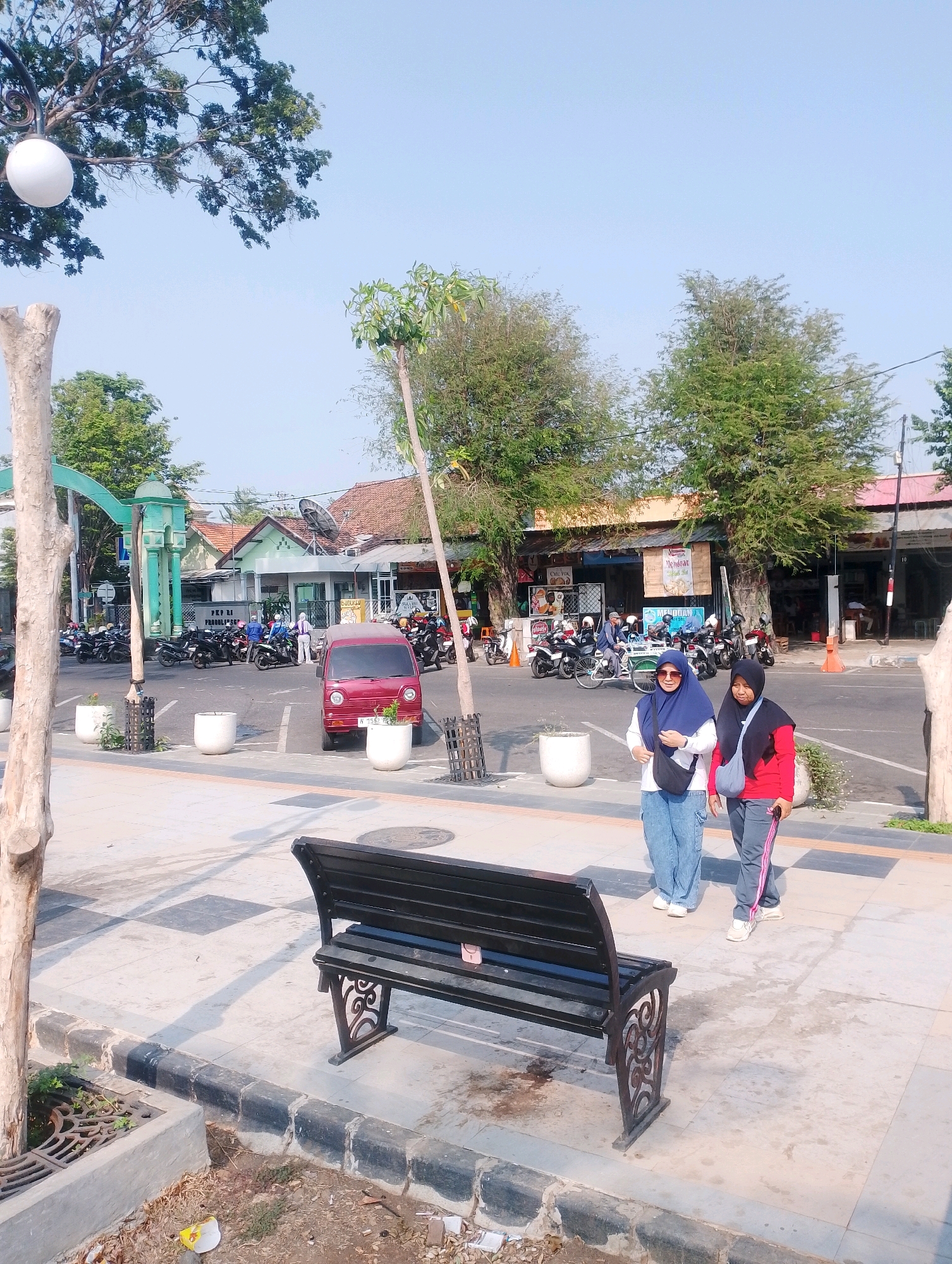 The image depicts an outdoor scene in the city of Probolinggo, Indonesia, likely a public area with a walkway and a street. In the foreground, a black metal bench with ornate legs sits on a paved area. To the left of the bench, a rough, light-colored tree trunk is visible.
Further back, two young women walk together on the paved walkway. The woman on the left wears a blue hijab, a white top, and light blue baggy pants. She carries a black crossbody bag. The woman on the right wears a black hijab and a red long-sleeved shirt with gray track pants that have a pink stripe. She carries a gray crossbody bag.
Beyond the walkway, a street is visible with parked motorcycles. A small red car is parked in a designated spot. Various buildings with tiled roofs line the street, including what appears to be a government building with a green archway entrance. Trees and foliage are scattered throughout the scene, suggesting a warm climate. The sky is clear and pale blue, indicating it is daytime. The overall impression is of a well-lit, possibly commercial or administrative area in an Indonesian city.
