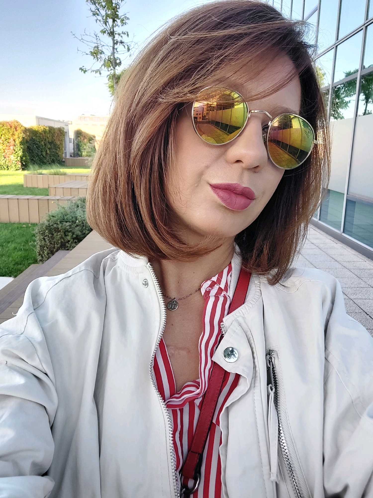 A woman with shoulder-length brown hair and mirrored round sunglasses takes a selfie outdoors. She is wearing a white bomber jacket over a red and white striped shirt. A thin silver necklace with a circular pendant is visible. Behind her, a modern building with reflective windows and a grassy area with tiered wooden seating are visible. The background suggests an urban park or a modern public space. The lighting indicates it is daytime. The scene appears to be in Sofia, Bulgaria.