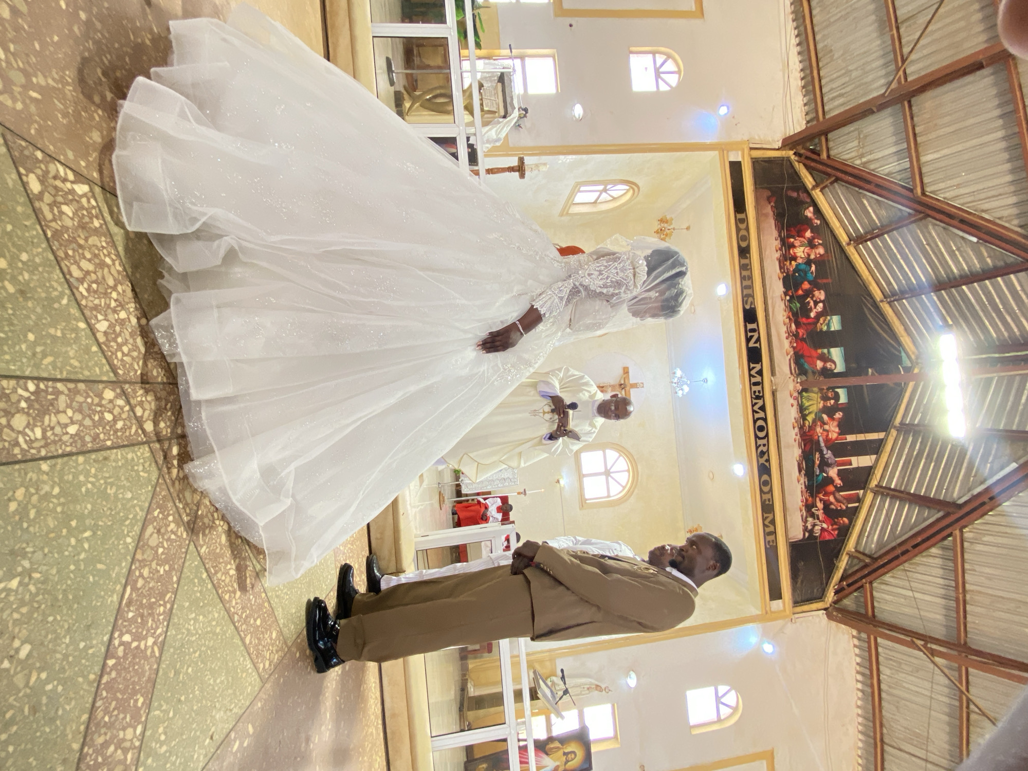 The image captures a wedding ceremony inside a church in Kuru Karama, Nigeria. The bride is wearing a white, long-sleeved wedding dress with a long veil. The groom, in a brown suit, stands next to her. An officiant stands behind them with a book. The church has arched windows, and a religious painting hangs on the wall. The floor is tiled in a geometric pattern.