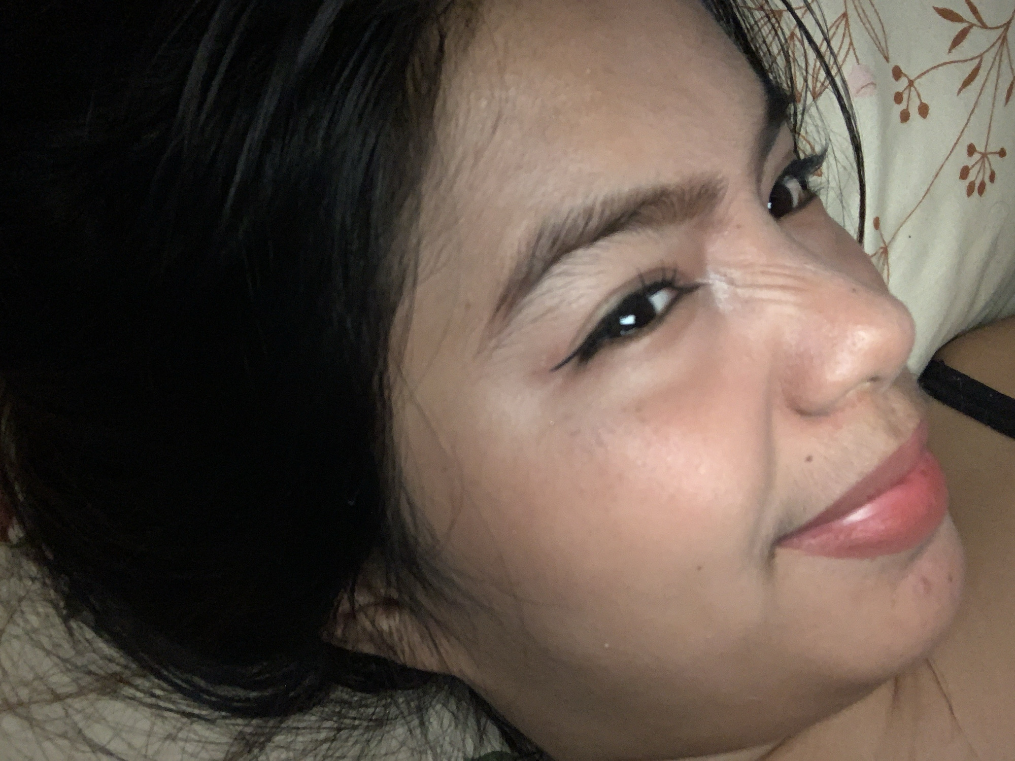 A close-up, angled shot captures a young woman's face in profile/three-quarters view, as she looks towards the camera with a relaxed expression and a slight smile. Her dark hair frames her face, appearing loose and flowing. She has dark eyes enhanced with winged eyeliner, and well-defined, dark eyebrows. Her lips are a glossy pink, and her skin tone is warm. Natural lines are visible on the bridge of her nose and around her eyes, consistent with movement or expression. Behind her head, a light-colored pillow with a subtle floral or leafy pattern is visible, suggesting an indoor setting, likely a bedroom. A dark strap, possibly from clothing, is visible on her right side. The lighting appears soft and indirect, possibly indicating an indoor environment at an unspecified time of day. No text is visible in the image.