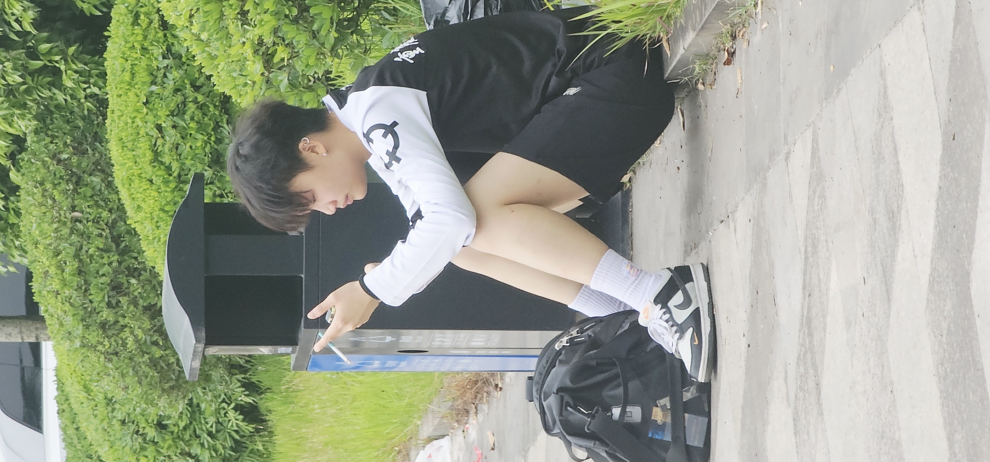 A person is squatting next to a public trash receptacle outdoors. The person is wearing a black and white long-sleeved shirt, black shorts, white socks, and black and white sneakers. They are holding a cigarette in their right hand, seemingly about to dispose of it in the receptacle. A black backpack is on the ground next to the person's feet. The setting appears to be an urban or suburban area, with greenery visible in the background. The location is Chongqing High-tech Zone, China. The walkway pavement has a distinctive rhombus pattern.