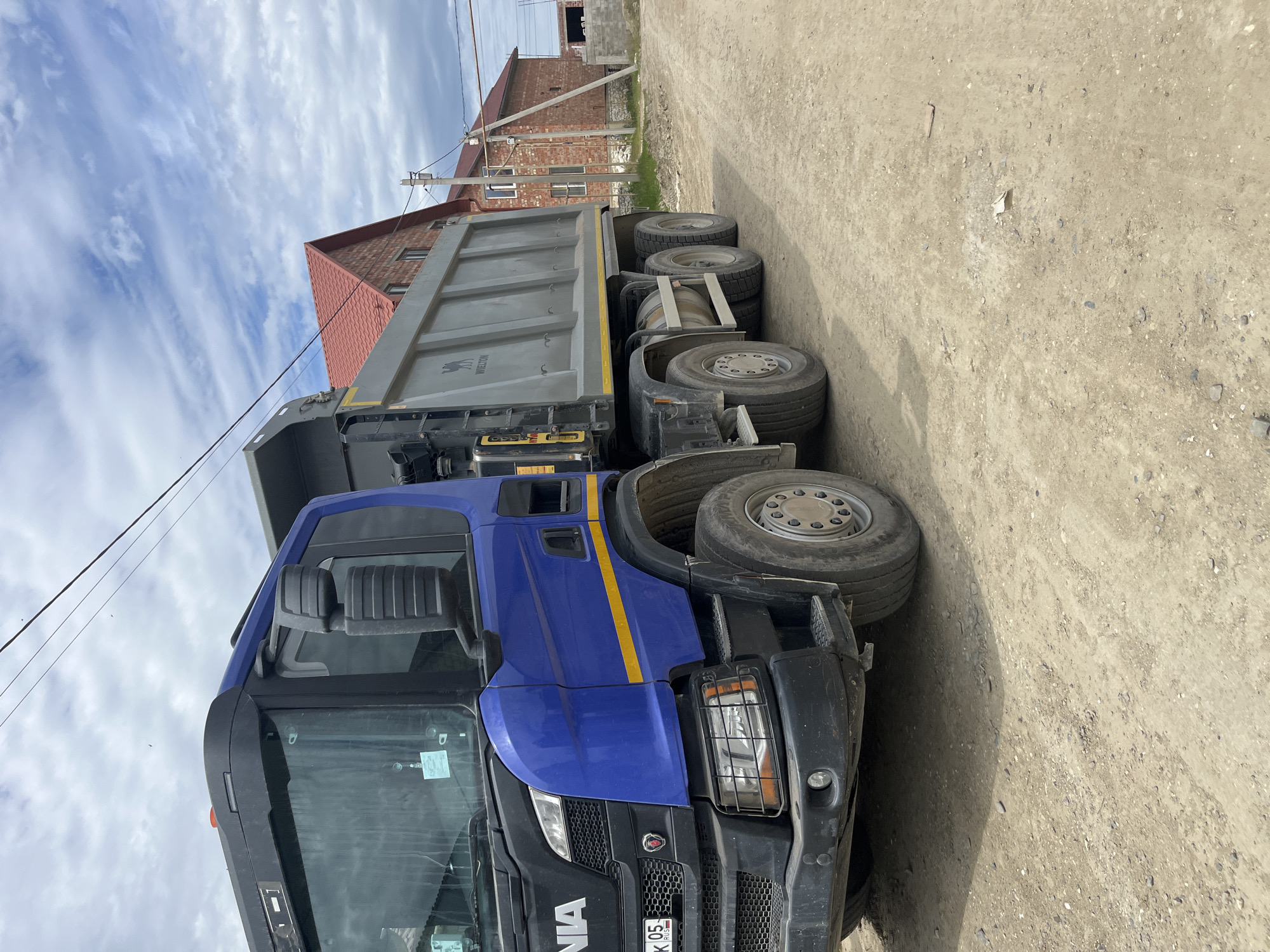 A blue and grey Scania dump truck is parked on a dirt road in Kaspiysk, Russia. A brick building is visible in the background. The truck's front is partially visible, including its license plate reading "AK 05 RUS". The sky is partly cloudy. The dirt road is uneven and pebbly.