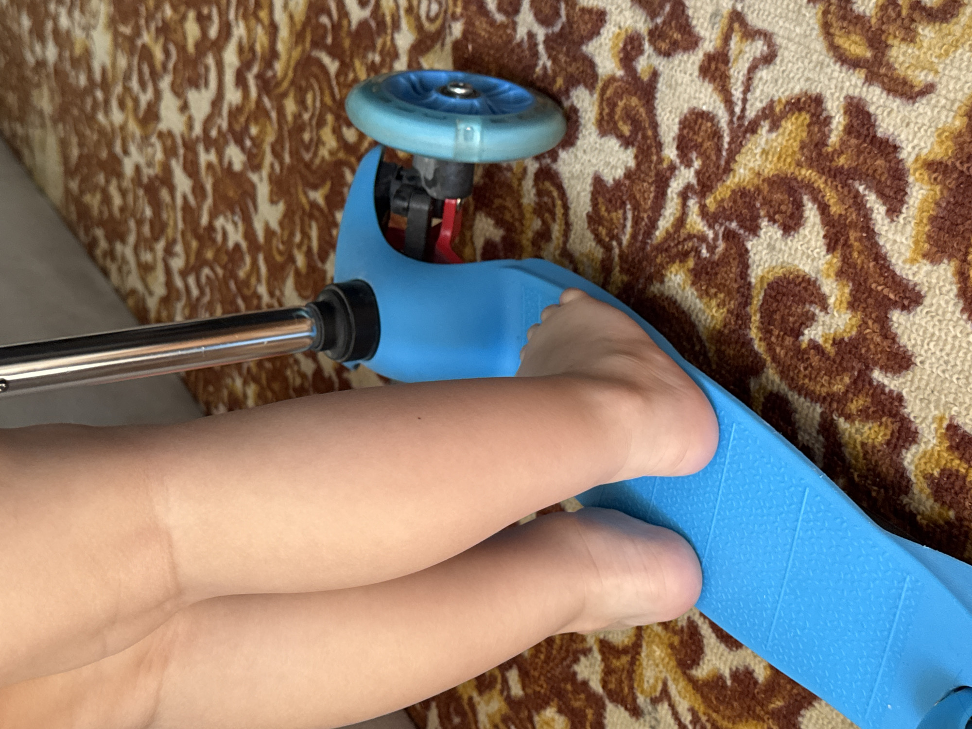 The image shows a close-up of a child's legs and feet resting on a blue scooter. The scooter has a blue wheel visible. The child's legs and feet are bare and appear to be resting on the scooter's deck. The background consists of a patterned carpet with brown and beige floral designs. The lighting suggests it is daytime, possibly indoors. The scene is focused on the child and the scooter, with no clear indication of location beyond the carpet pattern. No text is visible in the image.