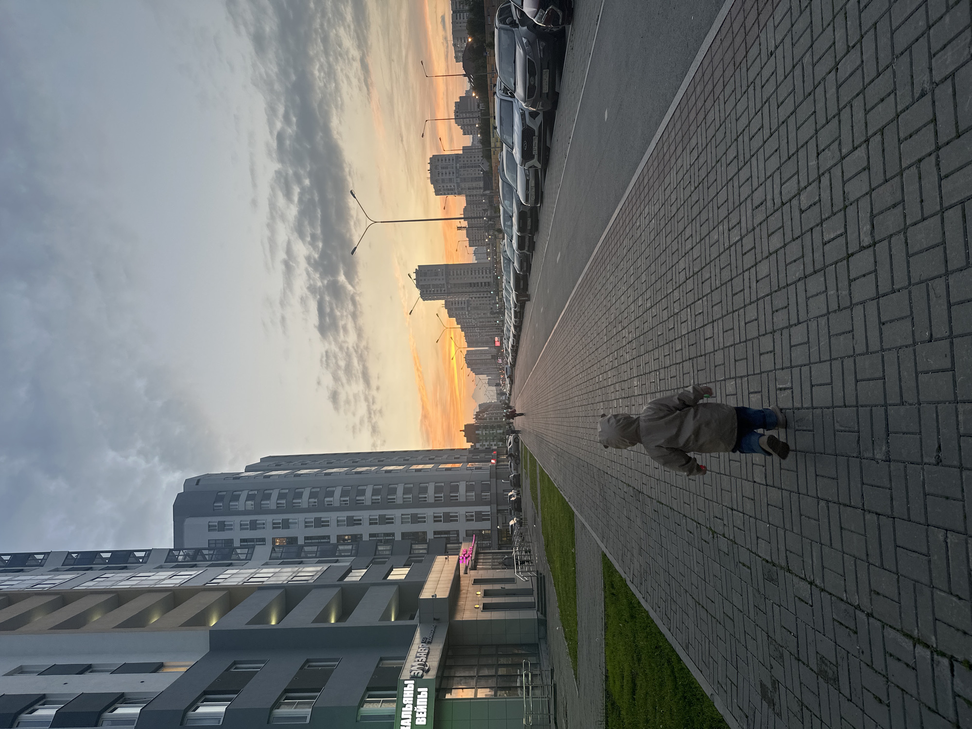In Yekaterinburg, Russia, at dusk, a young child wearing a hooded jacket and jeans walks away from the viewer down a brick-paved sidewalk. The child is in the foreground, centered on the path. To the left of the sidewalk are modern, multi-story residential buildings with numerous windows. To the right of the sidewalk is a street lined with parked cars, and beyond that, more multi-story buildings are visible in the distance. The sky is overcast with patches of bright orange and yellow, indicating the sunset. Streetlights are visible along the road. The scene appears to be an urban environment on a street in a residential district of the city.