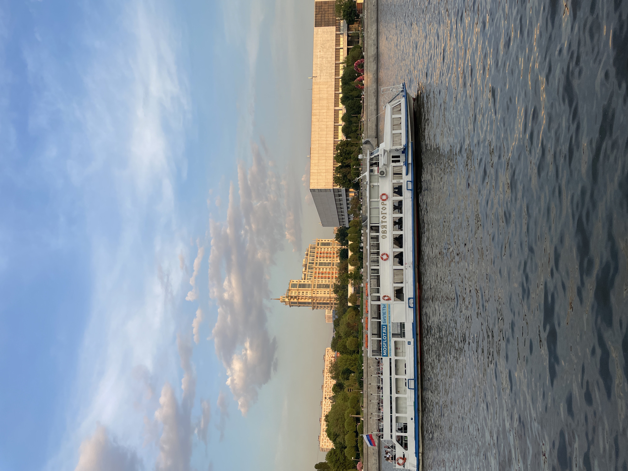 The image depicts a river scene in Moscow, Russia. A long, white passenger boat is docked parallel to a riverbank with a mix of modern buildings, green spaces, and mature trees. The boat has several decks and windows. A Russian flag is visible at the stern. The river water has a slightly rippled texture. The sky is blue with white clouds, suggesting either early morning or late afternoon light. The buildings along the riverbank are a mix of styles, from boxy concrete structures to more ornate apartment buildings.