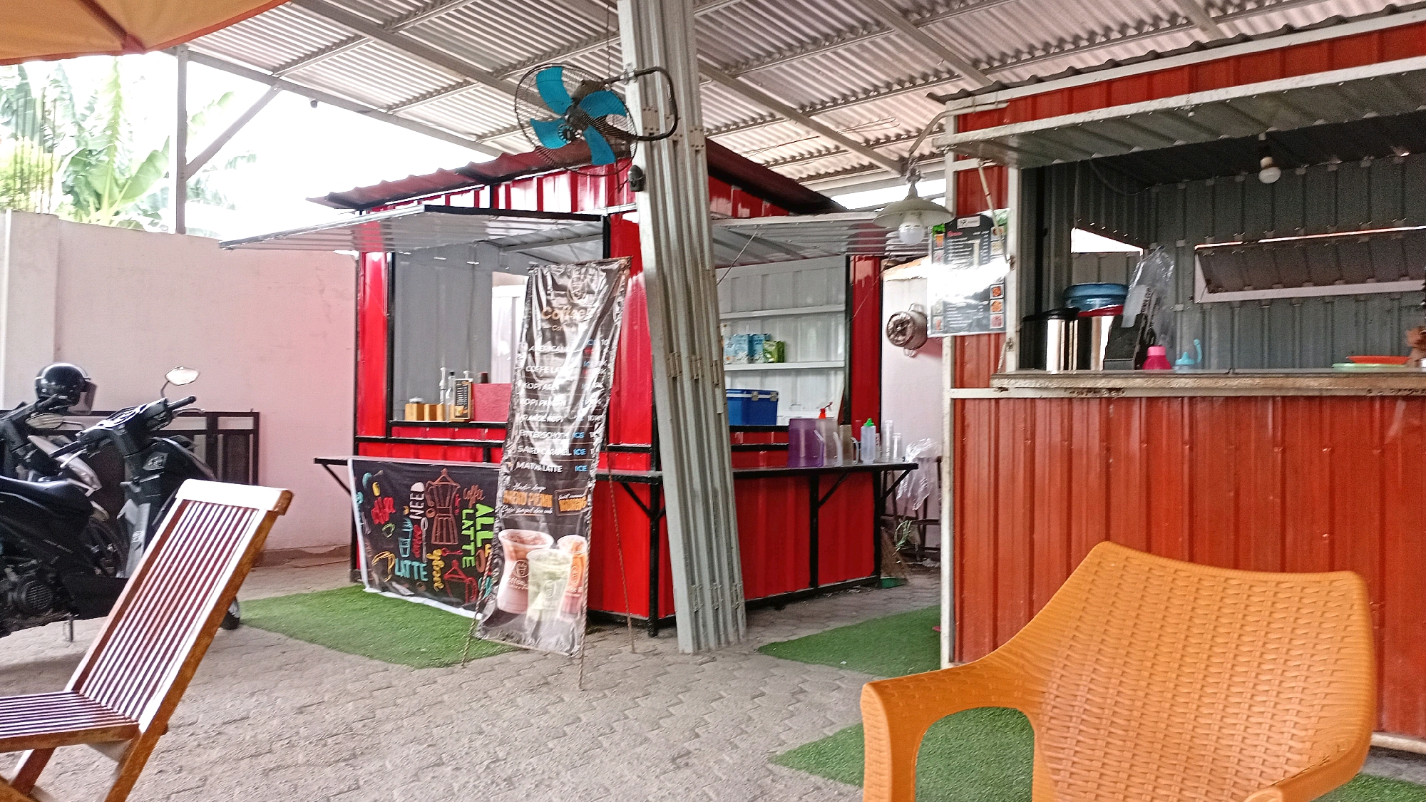 The image shows an outdoor seating area of a cafe, likely located in Jambi, Indonesia, given the presence of a moped which is a common mode of transportation. The cafe appears to be a small, open-air establishment with a corrugated metal roof supported by metal poles. The main structure is constructed from red corrugated metal panels, forming the walls of the serving area. There are two serving windows visible.
In the foreground to the left, a wooden slatted chair is positioned on a paved surface. To the left of the chair, a black moped with a helmet on its handlebars is parked. A large banner advertising coffee drinks, including "All Latte," is displayed vertically near the serving area, featuring images of coffee cups and brewing equipment. The floor is covered with artificial green turf in some areas.
In the background, a counter with various bottles and containers is set up behind the serving window. Shelving units are visible inside the serving area, holding stock and equipment. A blue fan is mounted on a metal pole overhead, and a simple pendant light fixture is also visible.
To the right of the image, another chair, this one made of orange woven plastic, is placed on the green turf. The overall scene suggests a casual and informal cafe setting, possibly catering to a local clientele. The environment is brightly lit by natural light filtering through the open roof structure.