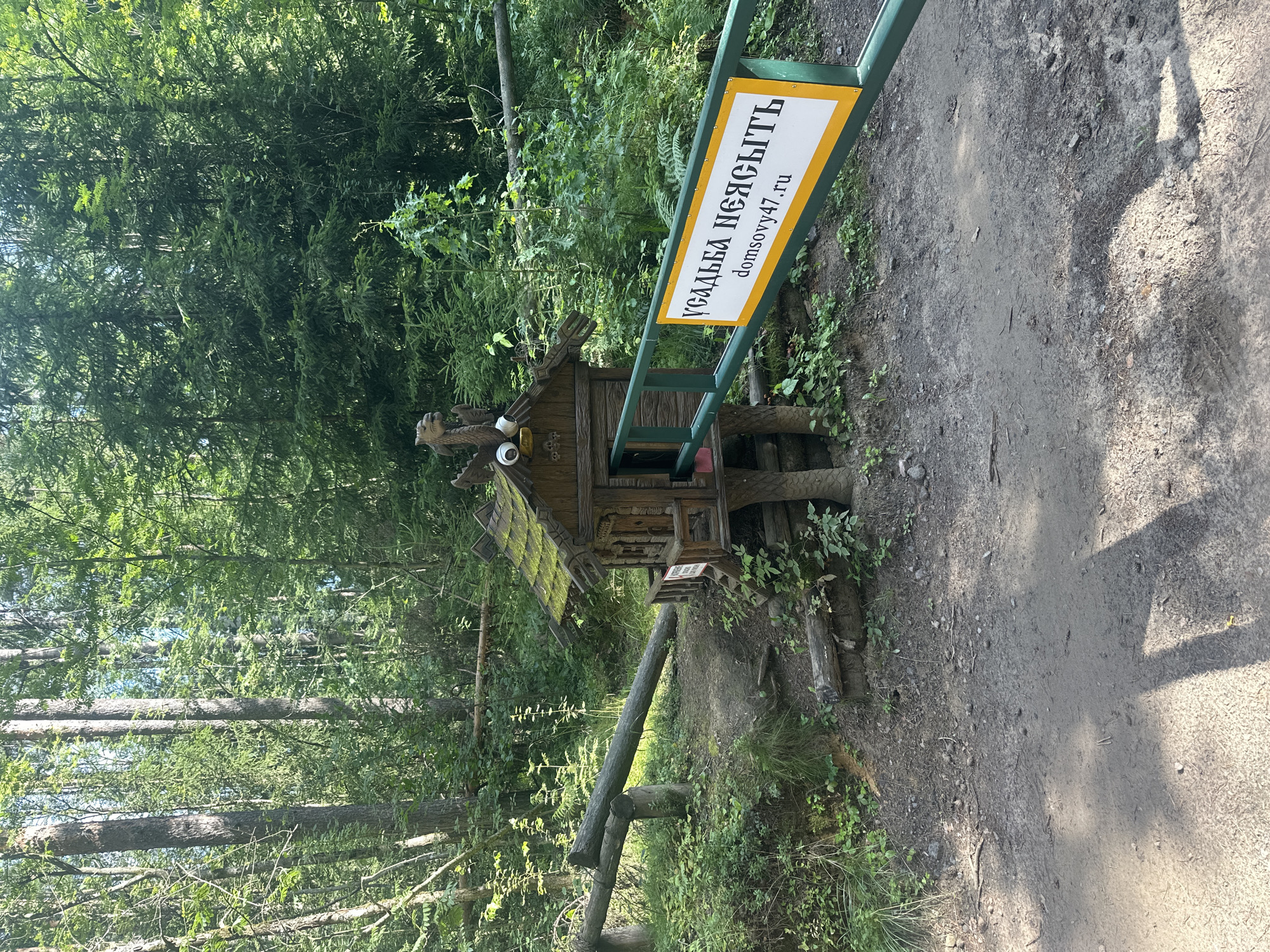 The image depicts a small, ornately carved wooden structure resembling a miniature house with a moss-covered roof, possibly a decorative element or a functional shed, situated in a wooded area. A green barrier gate with a white sign featuring yellow trim is in front of the structure. The sign displays Russian text and a website address: "УСАДЬБА НЕЯСЫТЬ domsovy47.ru". The surrounding environment is lush with green foliage, tall trees, and a dirt path leading towards the gate and the wooden structure. The scene appears to be outdoors, possibly at an entrance to a property in the village of Voeikovo, Russia, given the text on the sign.