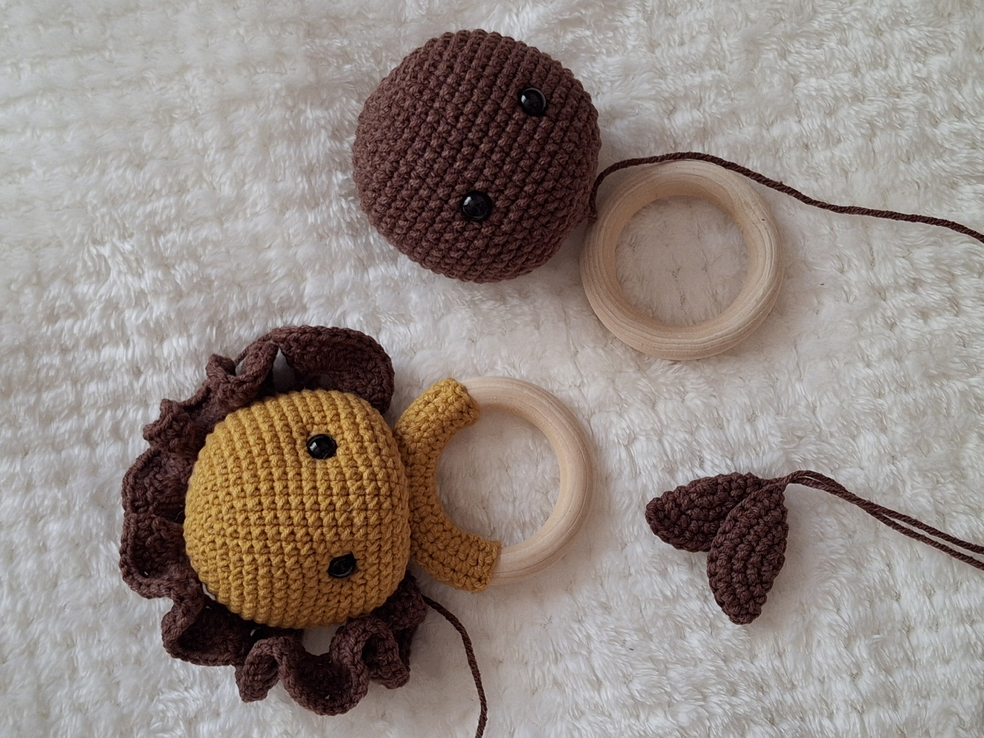 The image depicts a collection of crocheted baby toys on a soft, white, textured surface. There is a yellow and brown lion with a brown mane and a wooden ring attached. A brown sphere with black buttons is positioned near another wooden ring and a pair of brown ears with yarn strings. The setting is unspecified and there is no indication of it being in Chaikovsky, Russia.